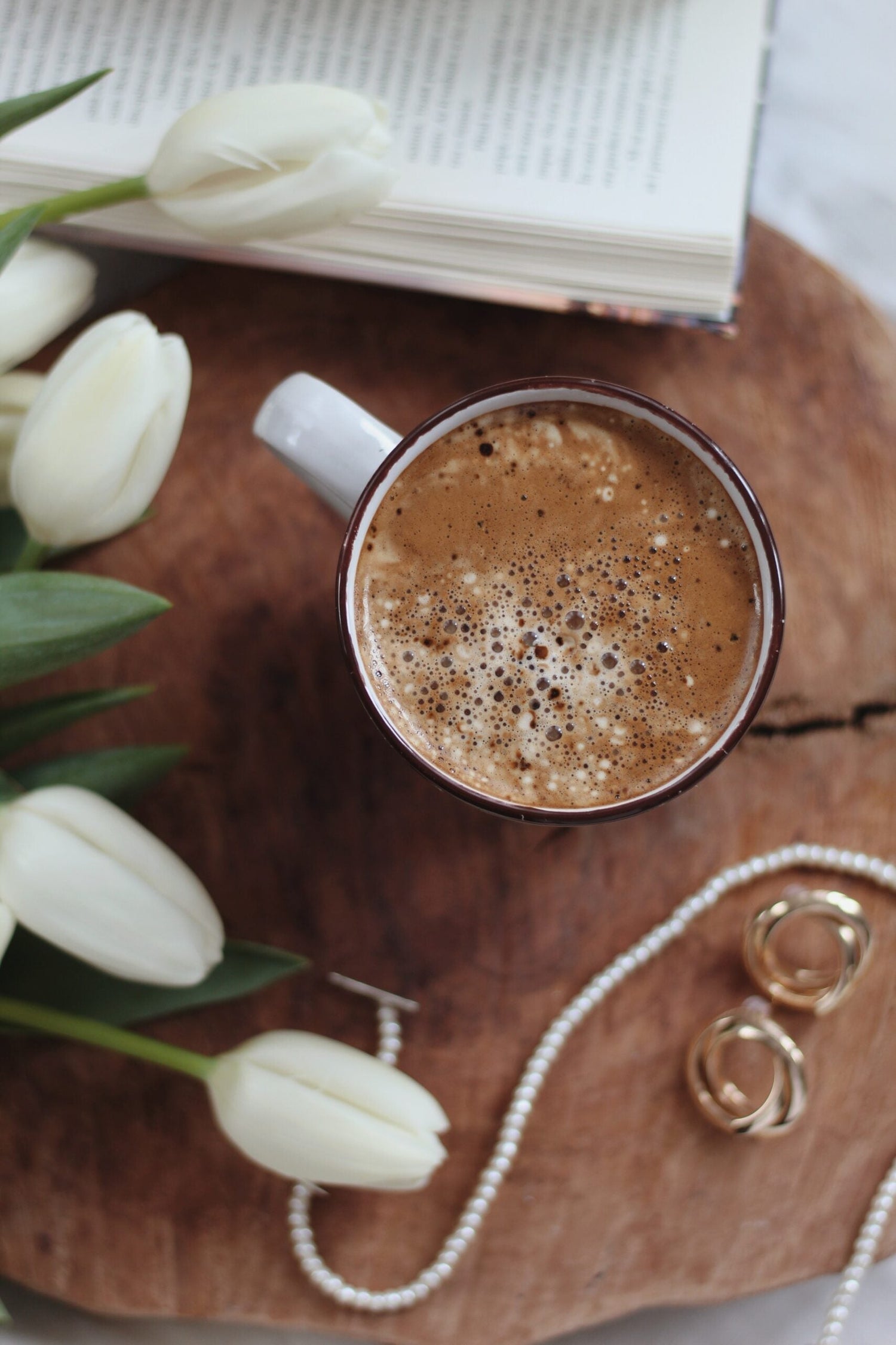 Top-down view of a frothy cacao mug on a wooden board with white tulips, an open book, and a pearl necklace