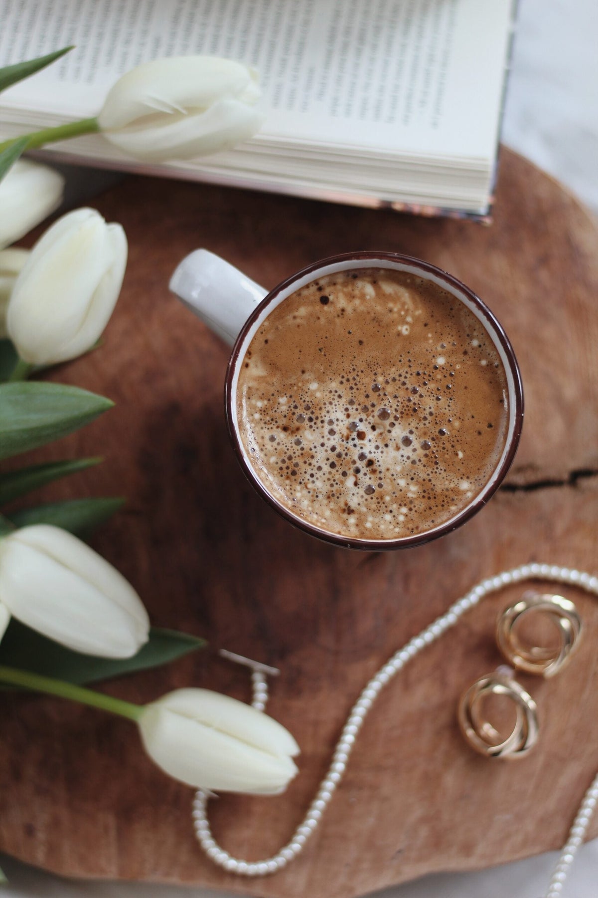 Top-down view of a frothy cacao mug on a wooden board with white tulips, an open book, and a pearl necklace