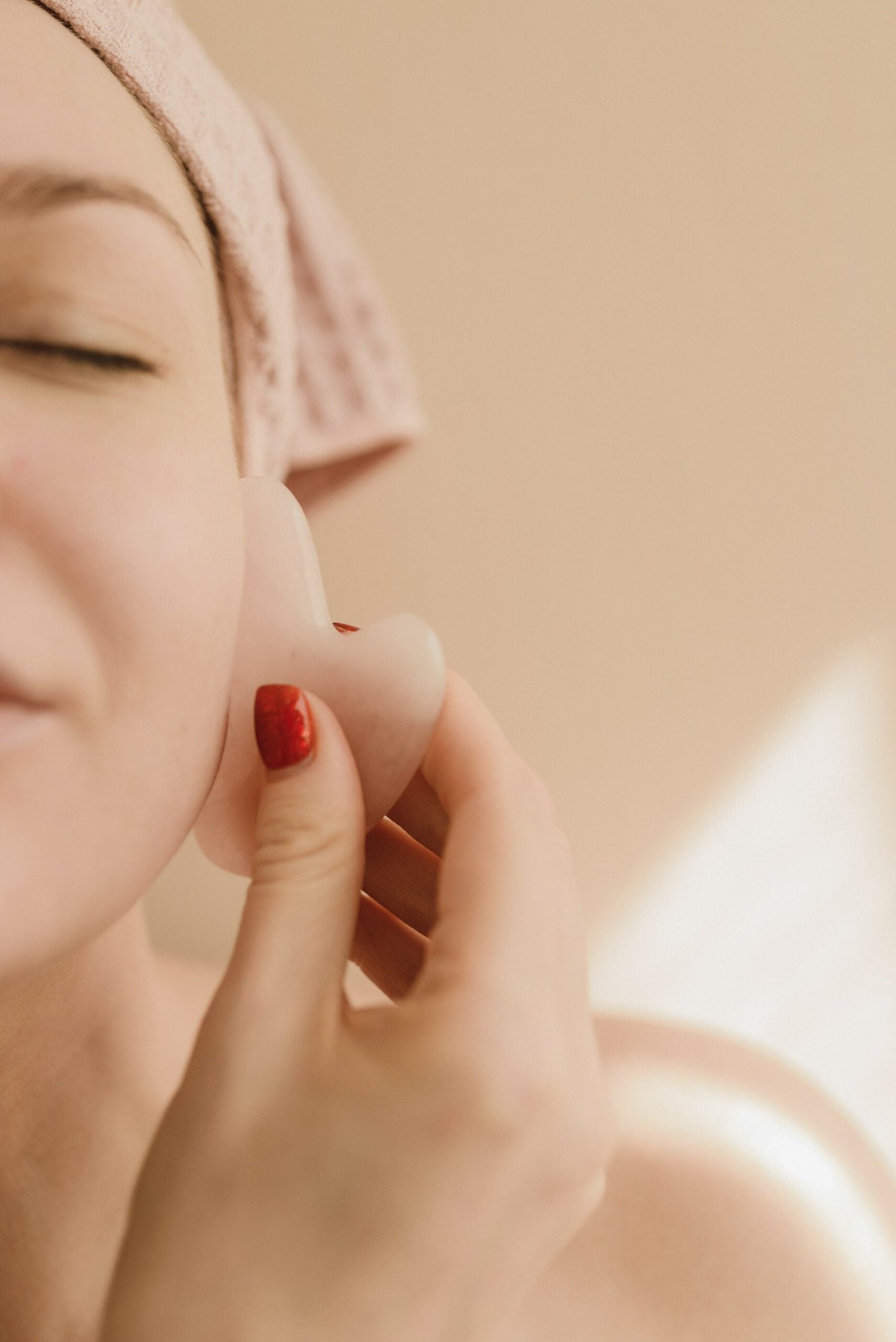 Woman using a pink gua sha tool on her jawline during a facial massage against a soft beige background