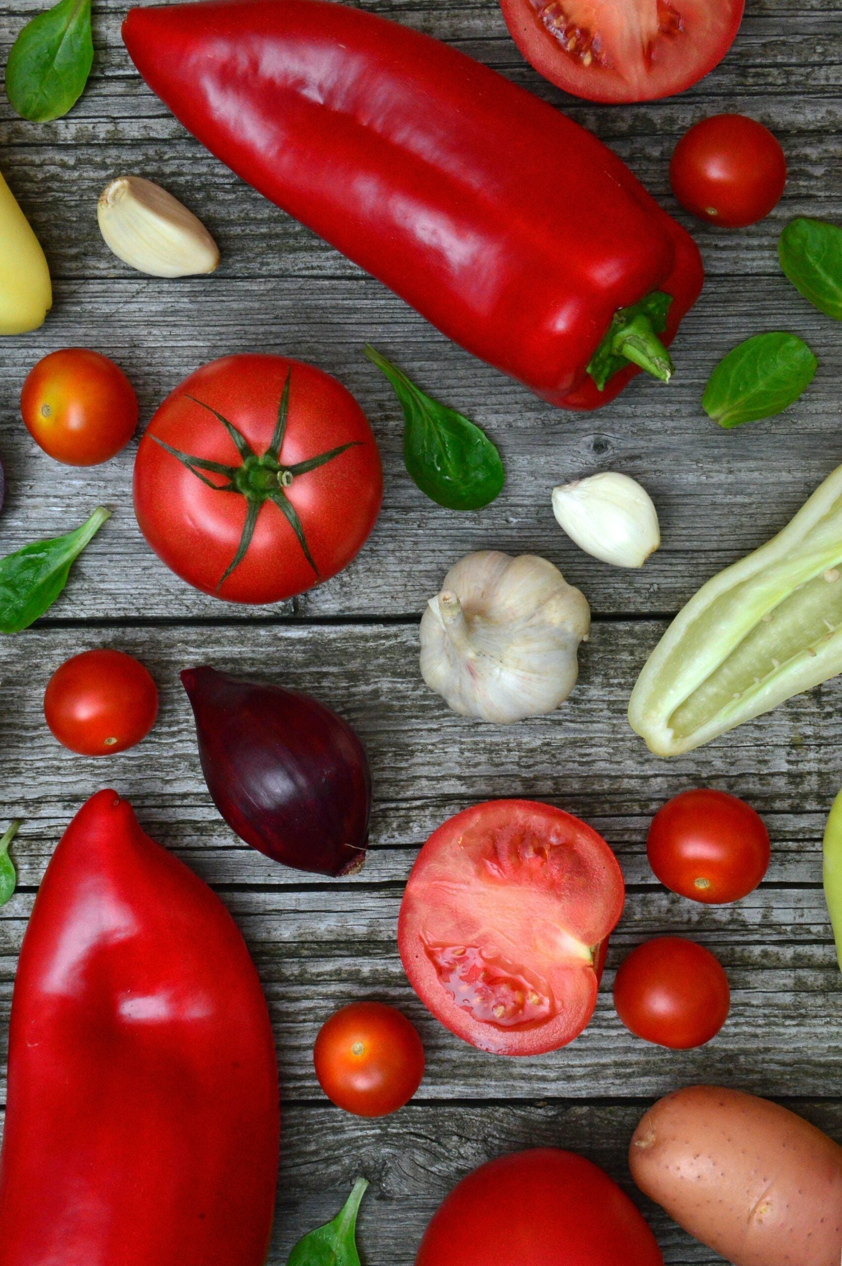 Red peppers, tomatoes, cherry tomatoes, garlic cloves, and basil leaves arranged on a weathered wooden board
