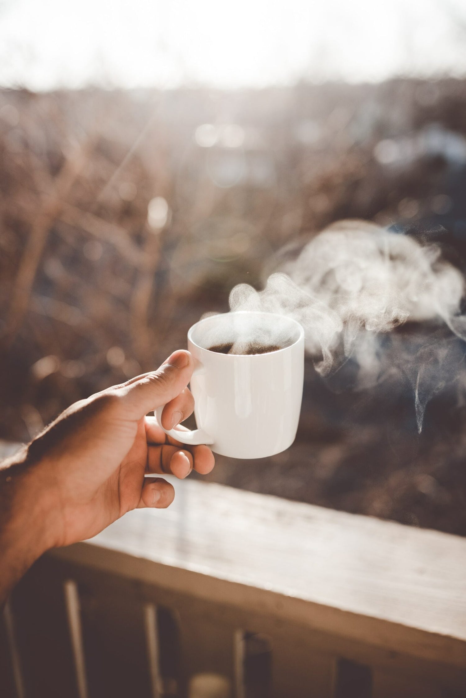 Hand holding a white mug of steaming coffee on a porch railing with a blurred outdoor background