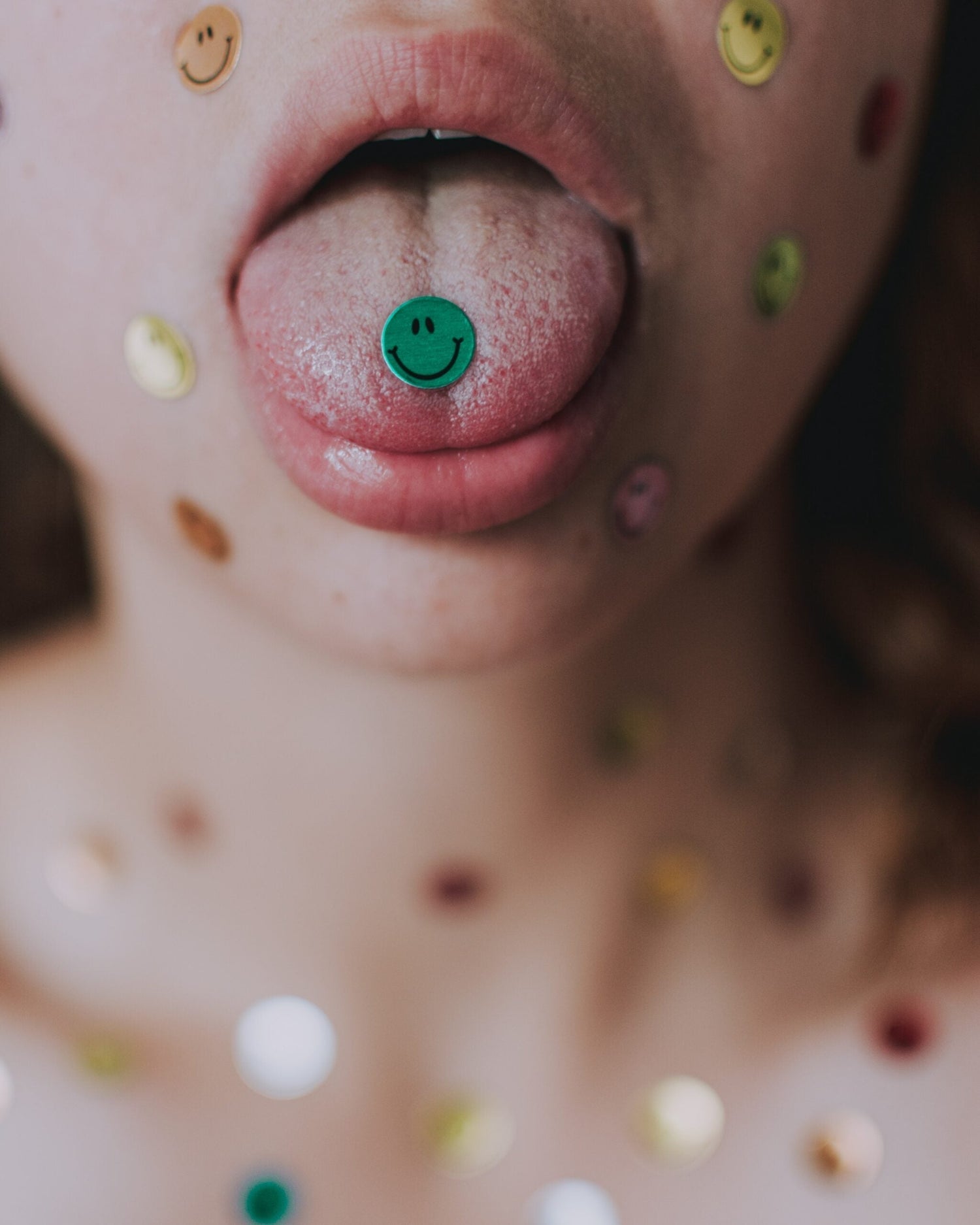 Close up of a person’s tongue with a green smiley sticker on the tip and colorful smiley stickers on the face and chest