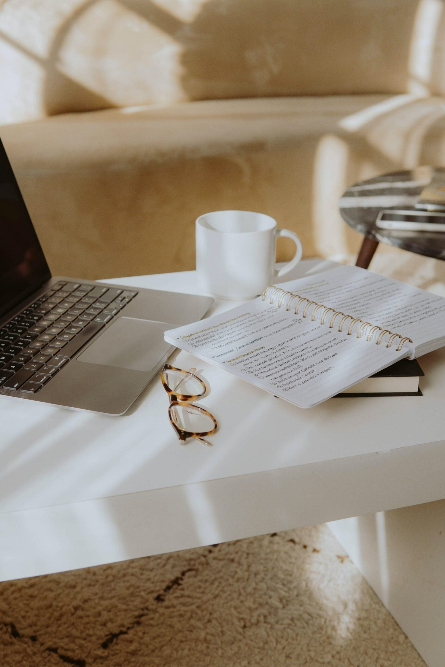 Sunlit workspace with open laptop, spiral notebook of handwritten notes, tortoiseshell glasses, and white coffee mug on a white table