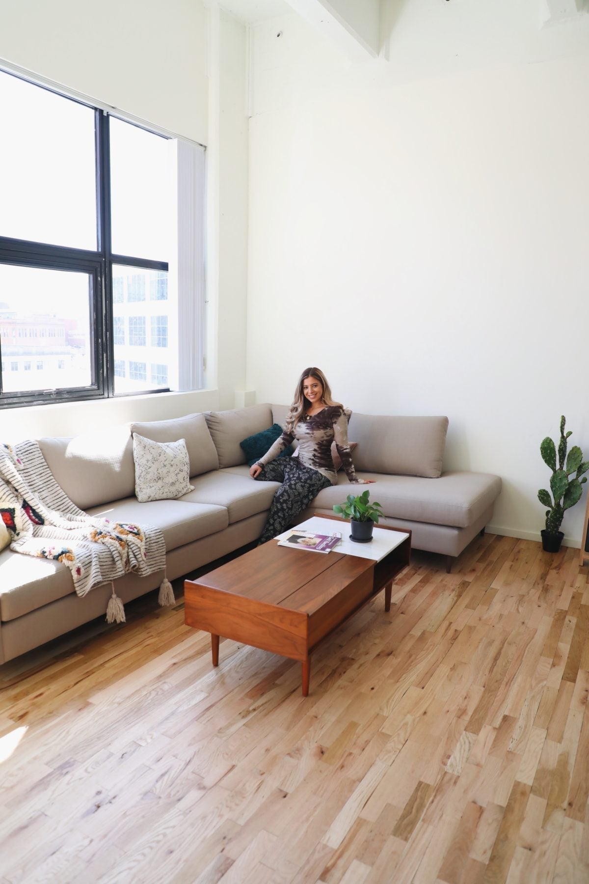 Woman sitting on a light beige corner sectional couch in a bright loft living room with a wooden coffee table and potted plants