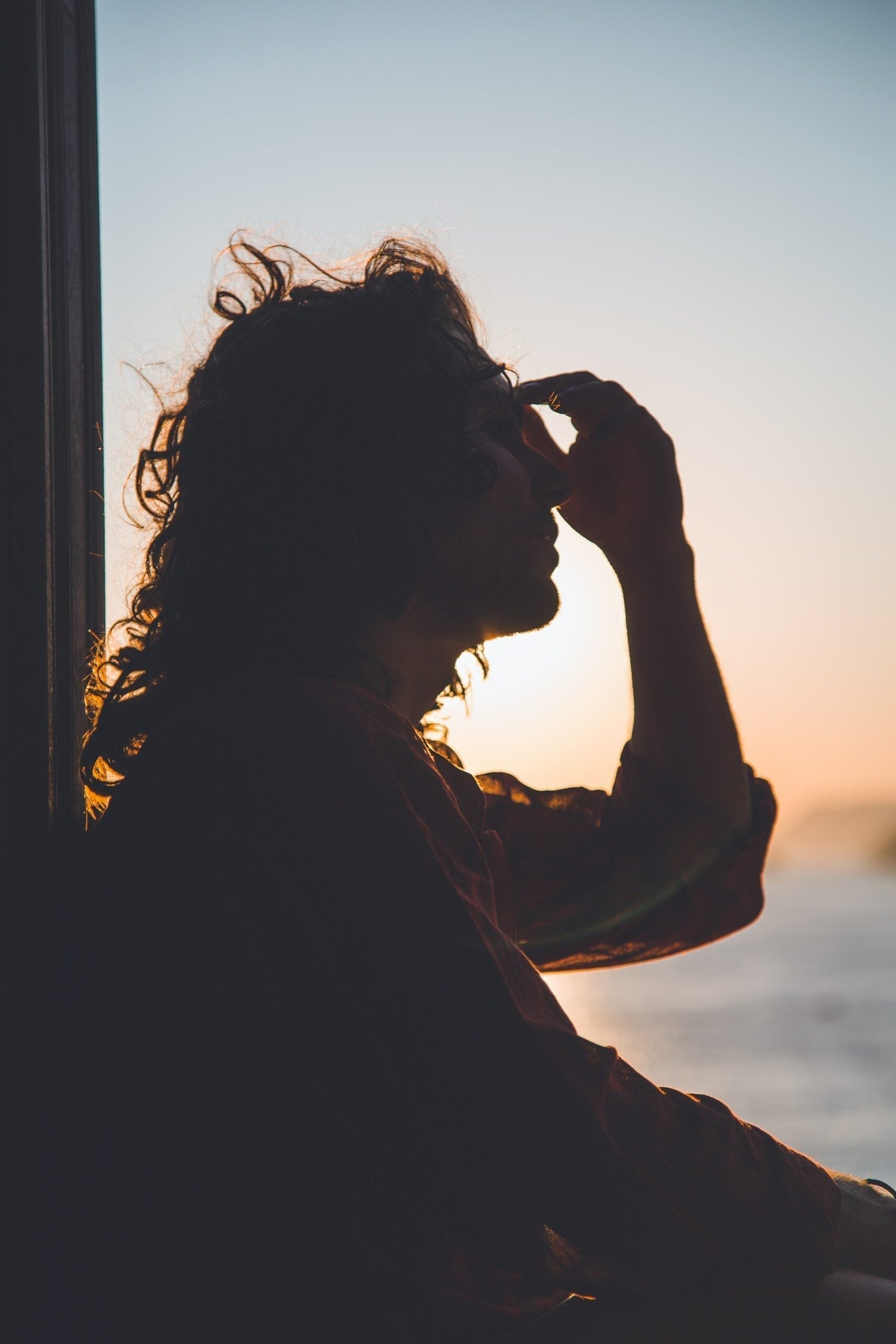 Silhouette of a person holding their forehead at sunset by the water, suggesting fatigue and contemplation