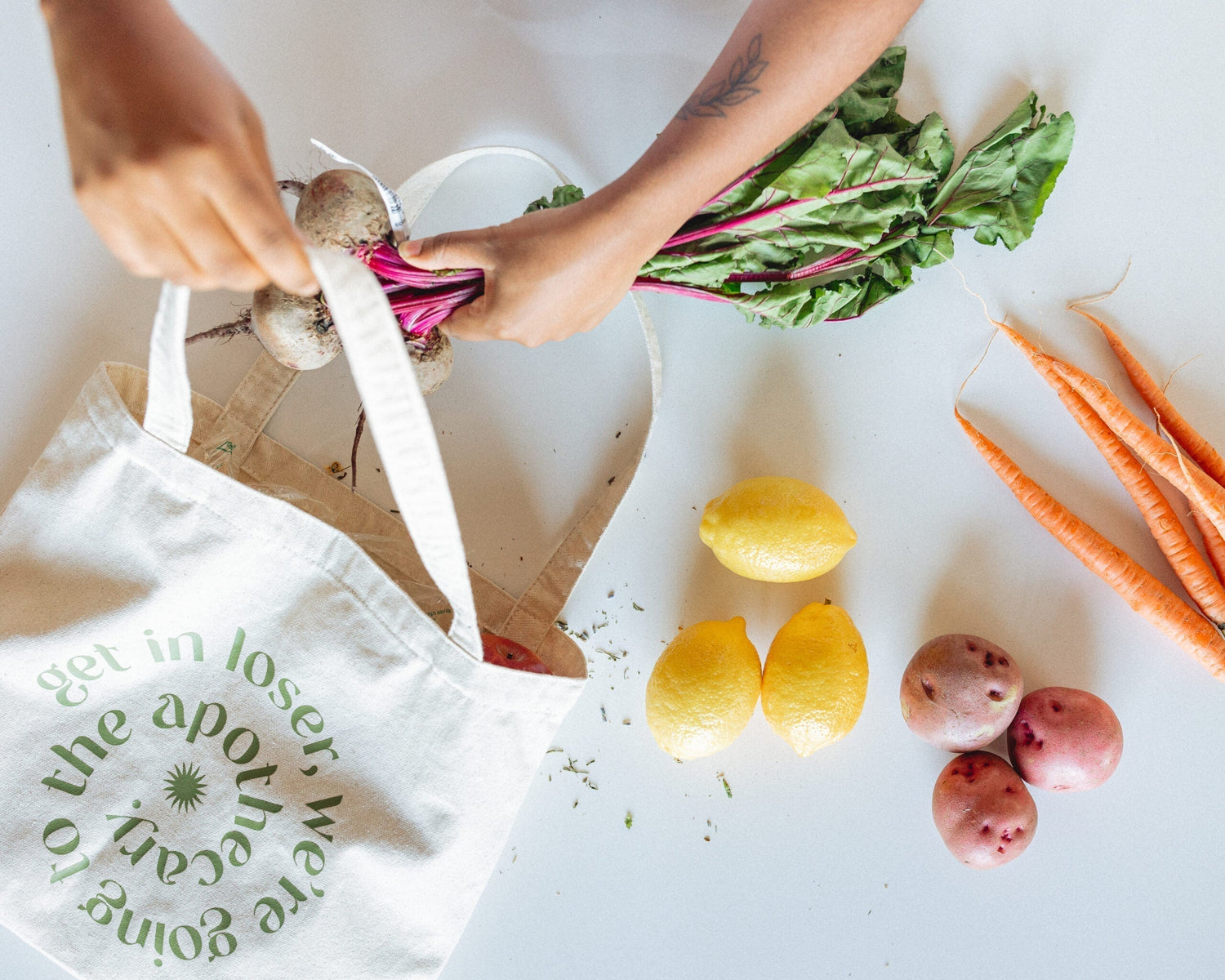 Hands placing a bunch of beets into a canvas market tote beside lemons, carrots, and red potatoes on a white table