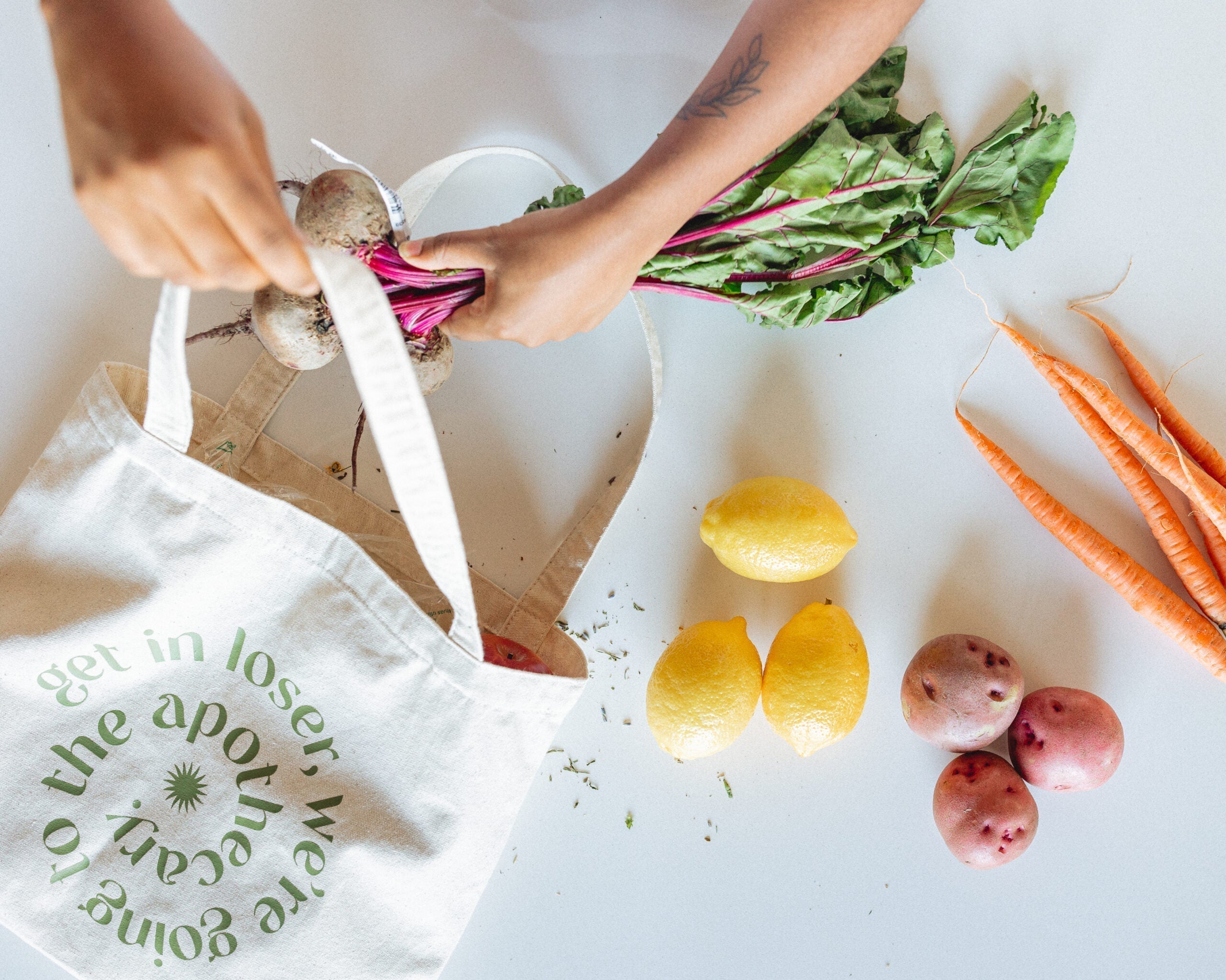 Hands placing a bunch of beets into a canvas market tote beside lemons, carrots, and red potatoes on a white table
