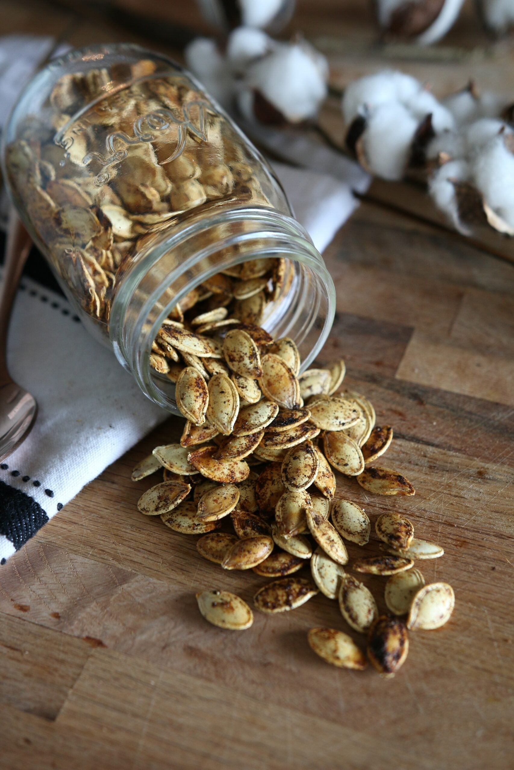 Close-up of roasted pumpkin seeds spilling from a tipped glass jar, seasoned with spices on a wooden cutting board with cotton bolls in the background
