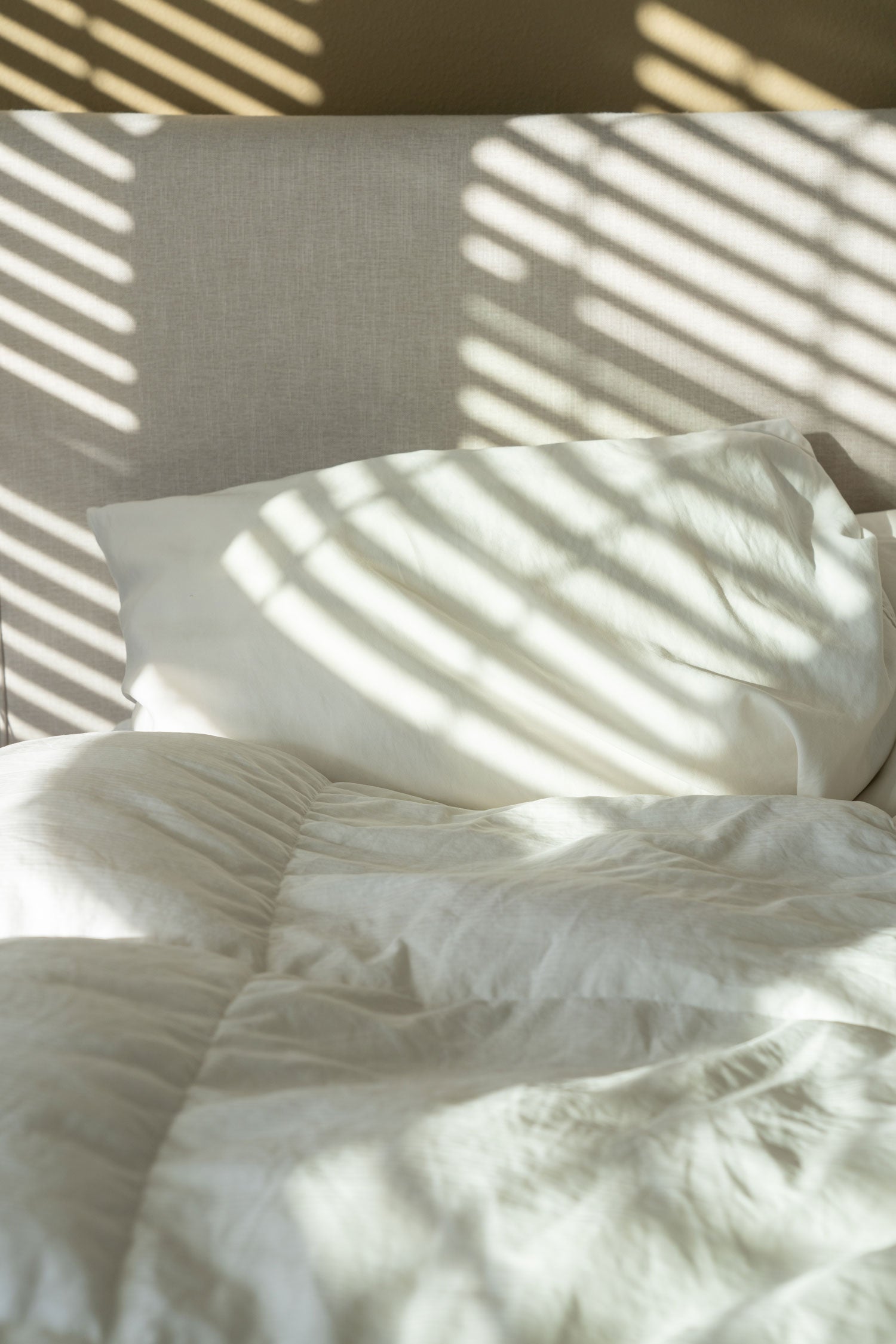 Sunlit bedroom pillow and rumpled white bedding with diagonal window-blind shadows