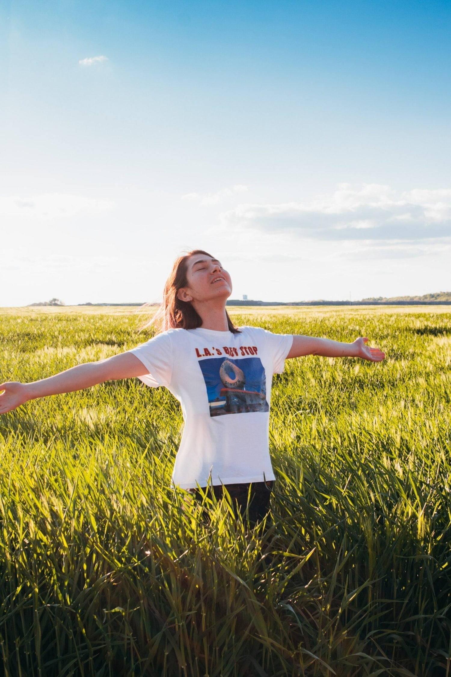 Young woman standing in a sunlit grassy field with her arms outstretched and eyes closed under a blue sky
