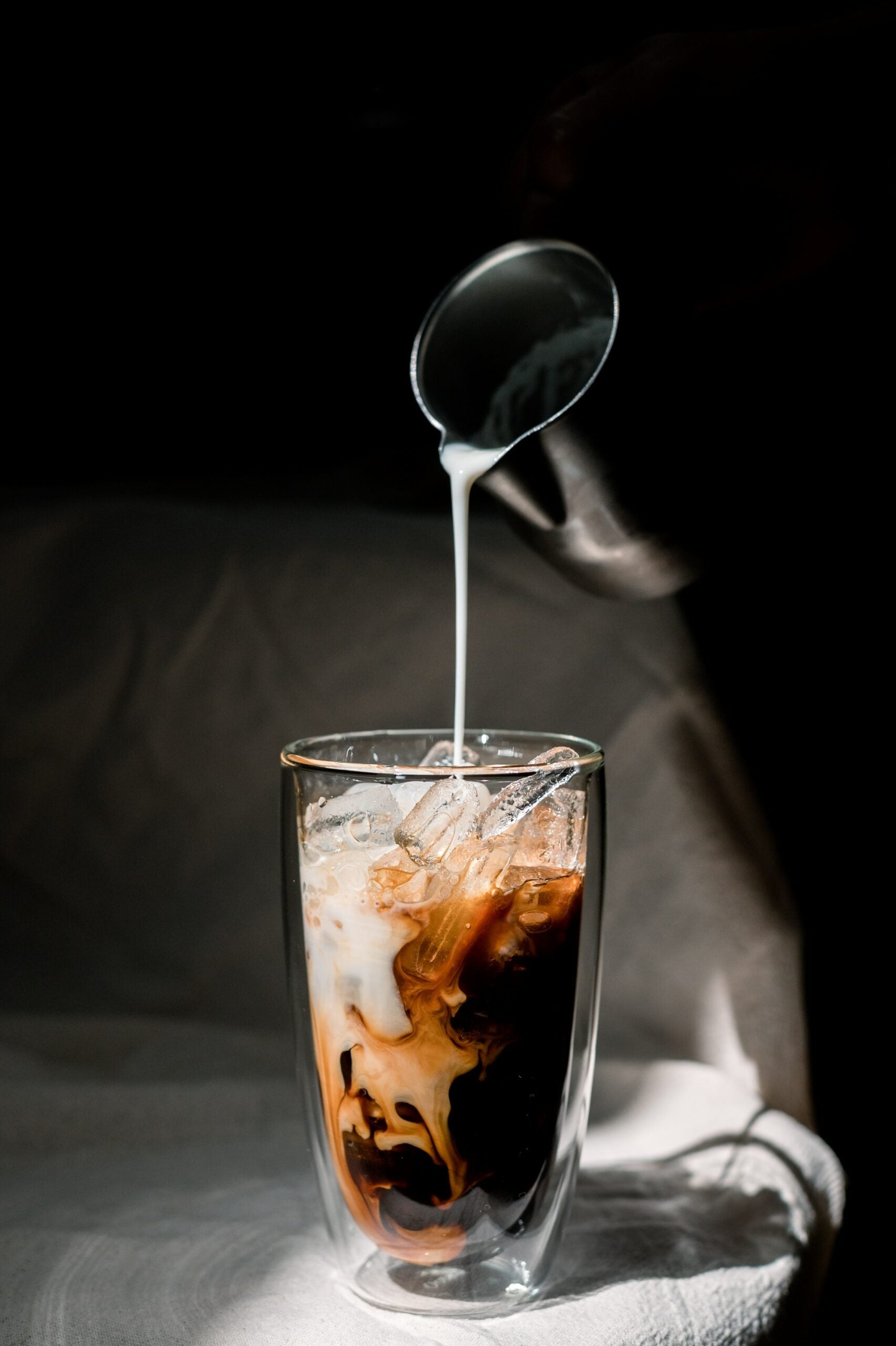 Hand pouring dairy-free milk from a spoon into a tall glass of iced coffee with ice against a dark background