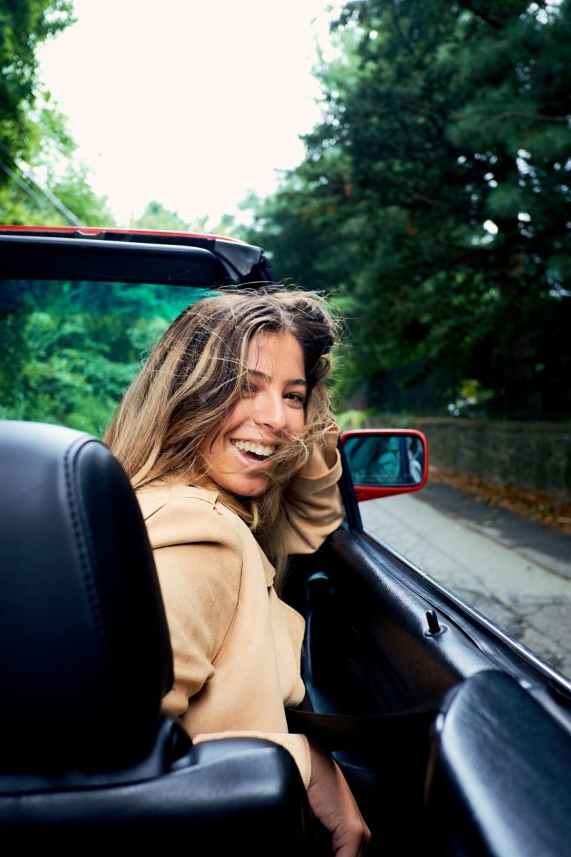 Young woman smiling and looking back while sitting in the passenger seat of a red convertible on a tree-lined road