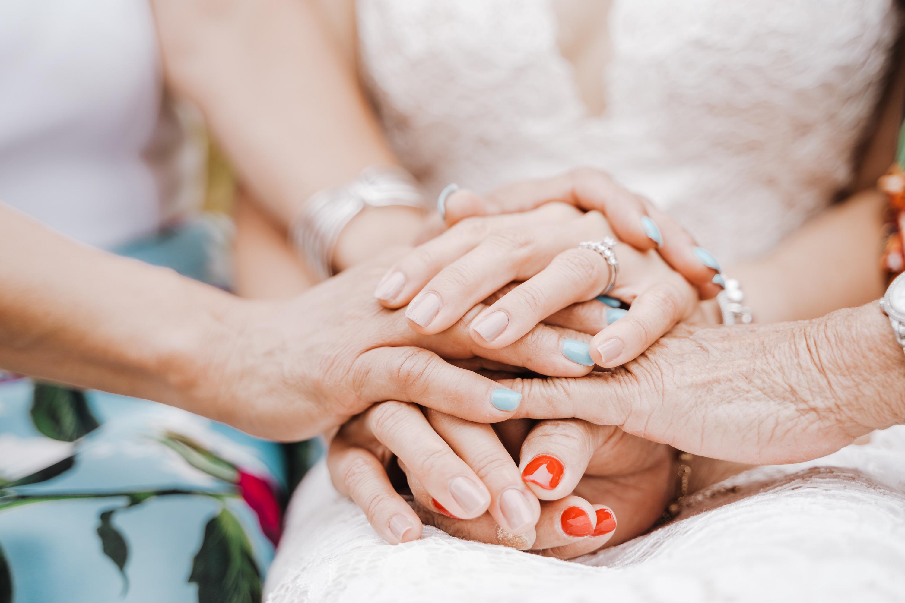 Multiple women’s hands of different ages stacked in a supportive gesture, some wearing rings and colorful nail polish over a white lace dress