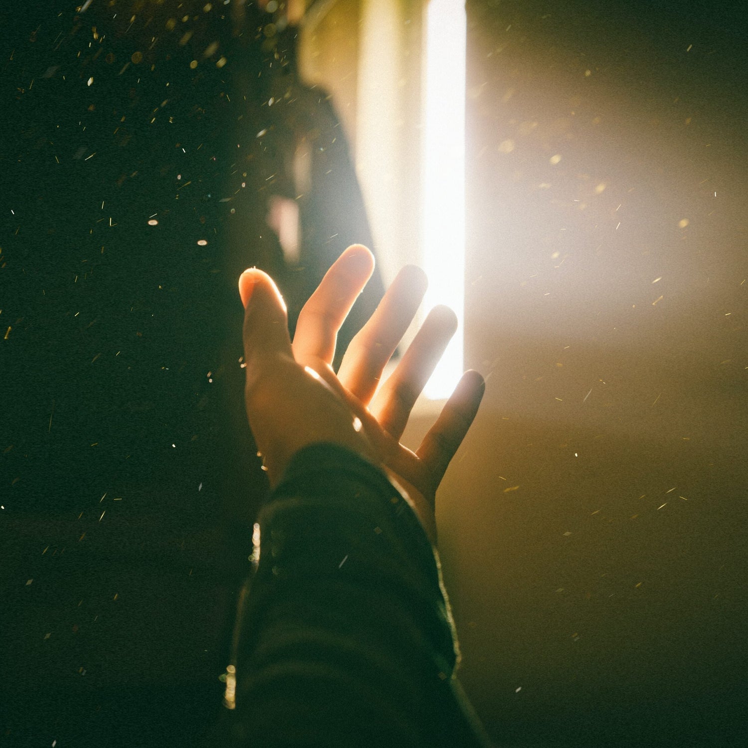 Hand reaching toward bright window light with floating dust particles in a dim, warm room