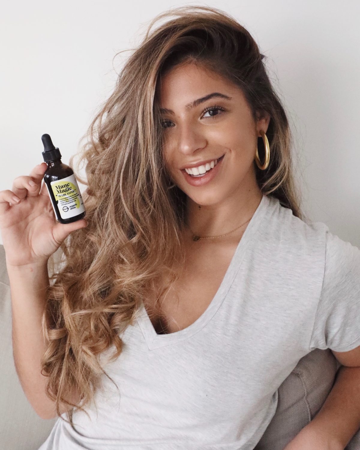 Woman smiling and holding a bottle of Mane Magic hair tonic while seated against a white wall
