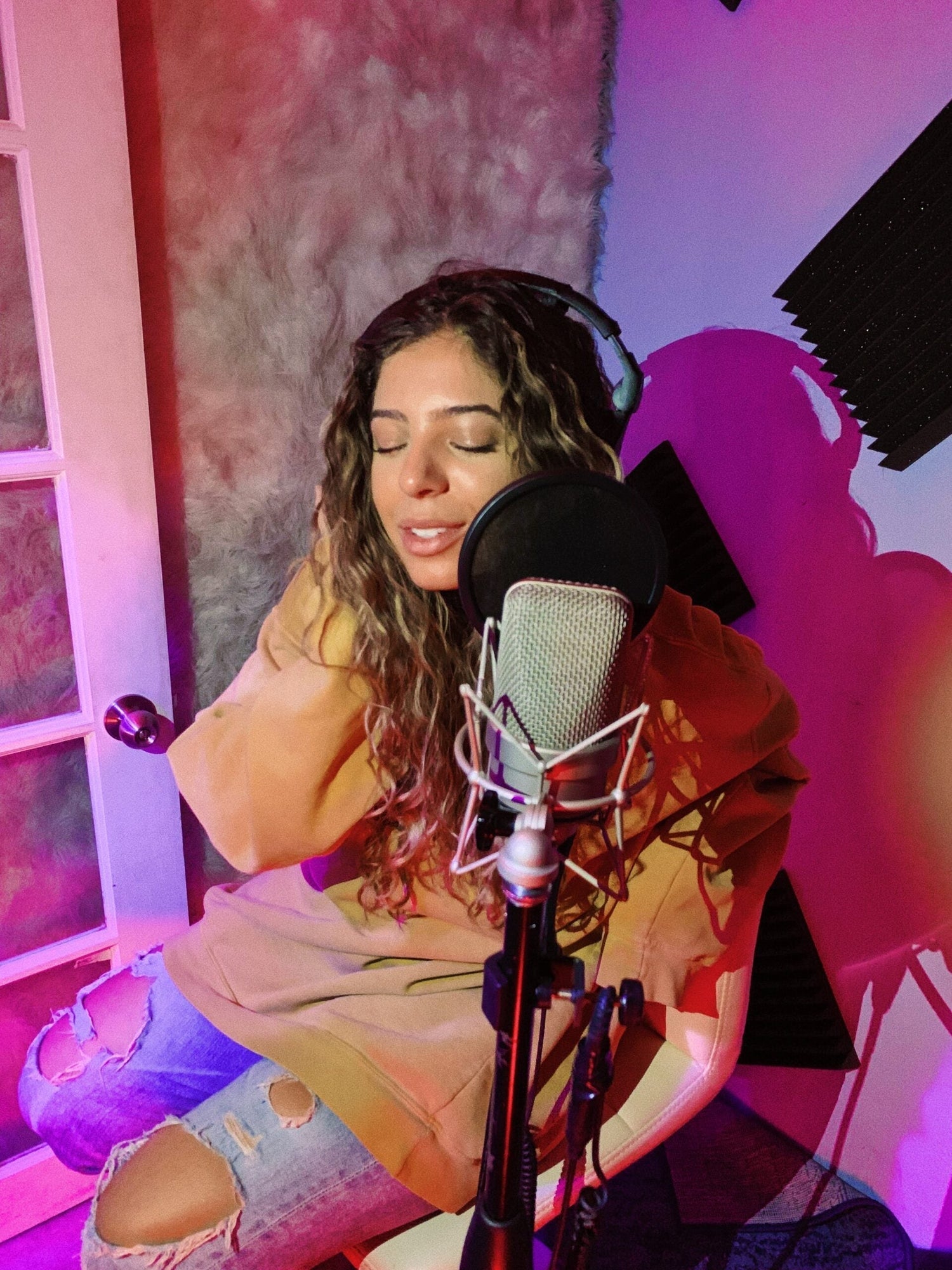 Woman with eyes closed singing into a studio microphone while wearing headphones in a colorful recording booth