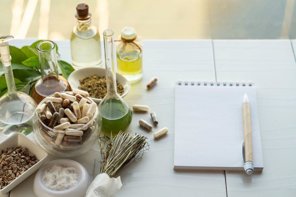 Herbal remedies scene with a bowl of capsules, glass tincture bottles, dried herbs and a blank notepad on a sunlit table