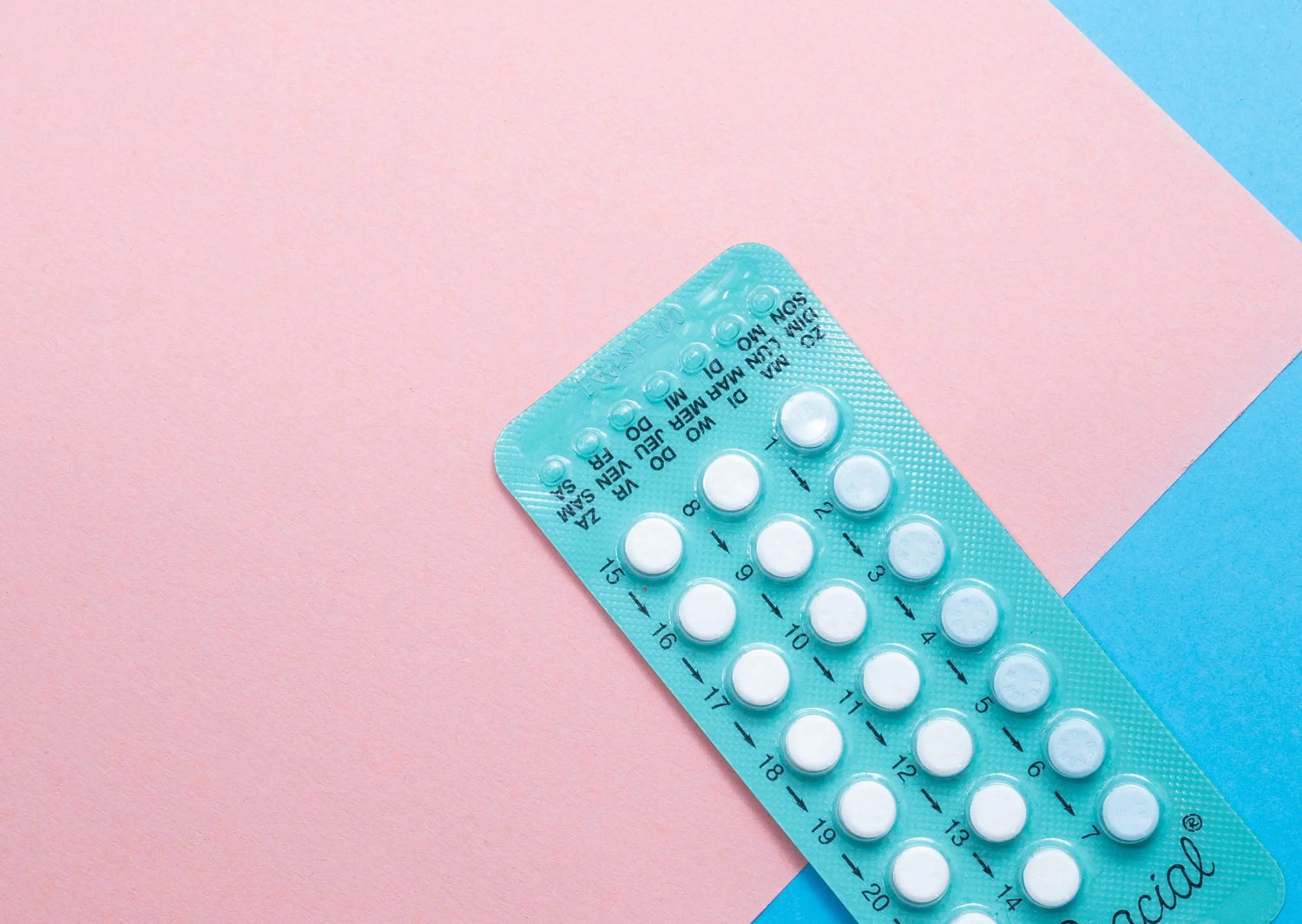 Angled top-right view of a teal blister pack of birth control pills showing numbered daily tablets on a pink and blue paper background