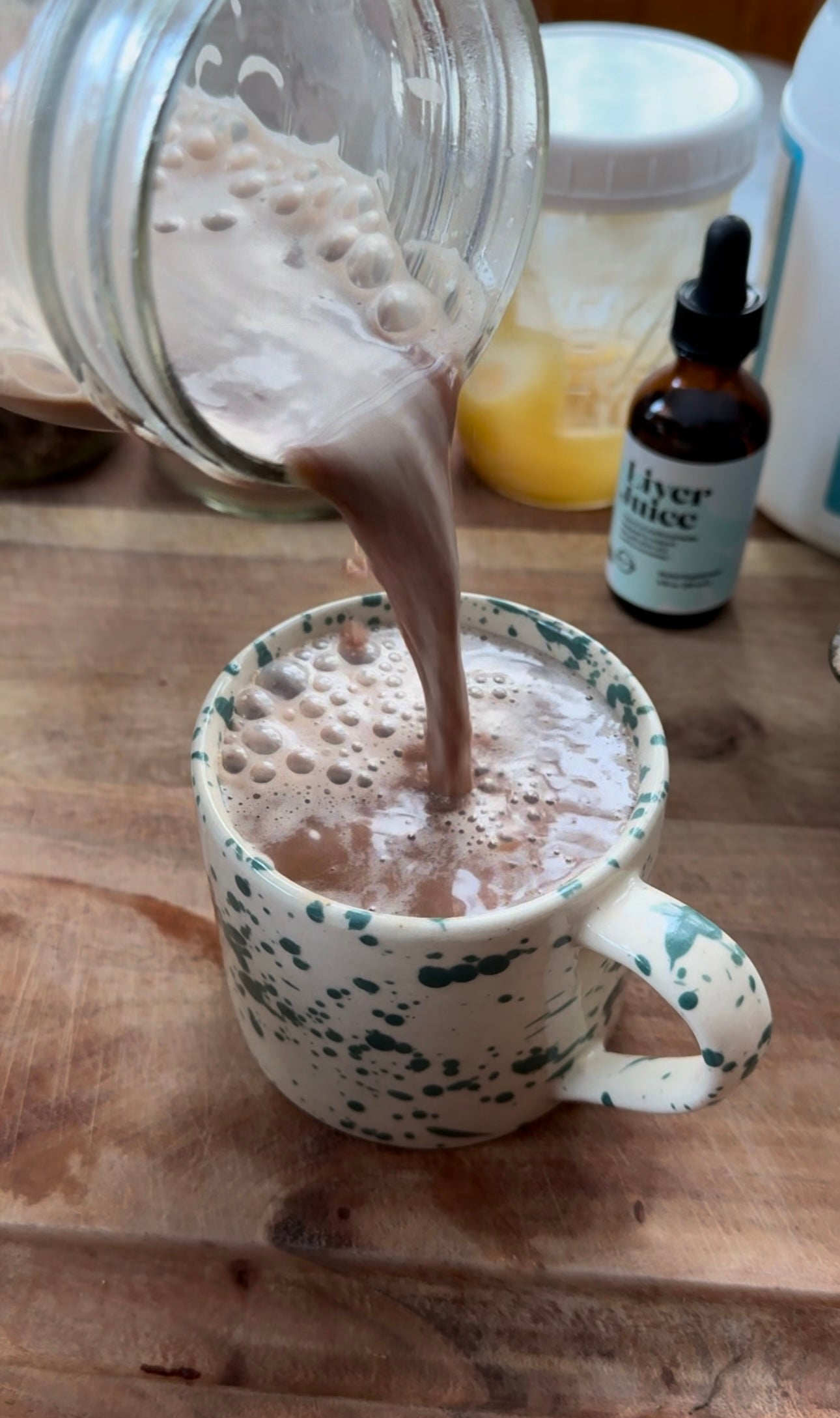 Jar pouring frothy dandelion latte into a green-speckled mug on a wooden counter, jars and a dropper bottle blurred behind