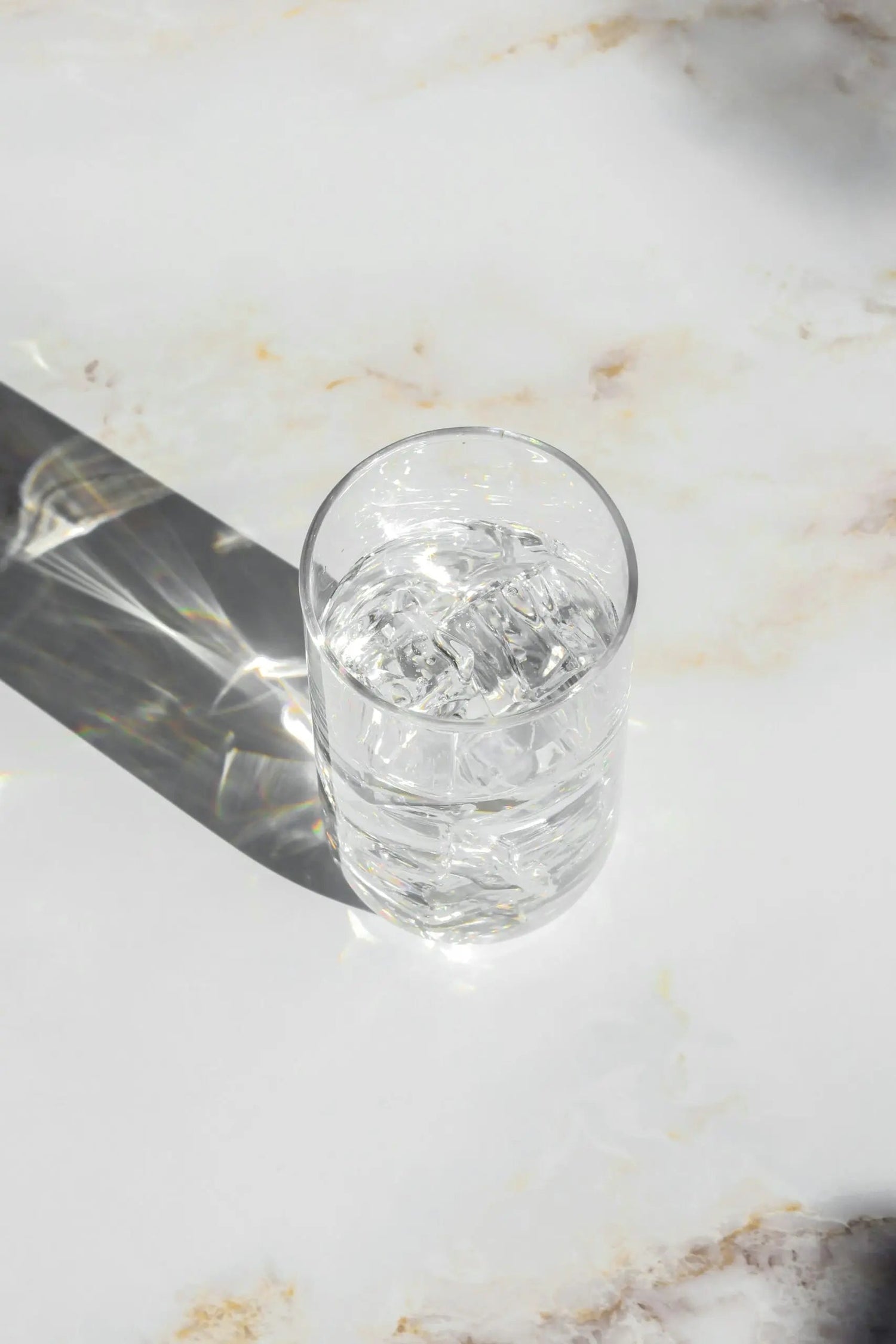 Overhead view of a glass of iced water casting a long prismatic shadow on a marble countertop