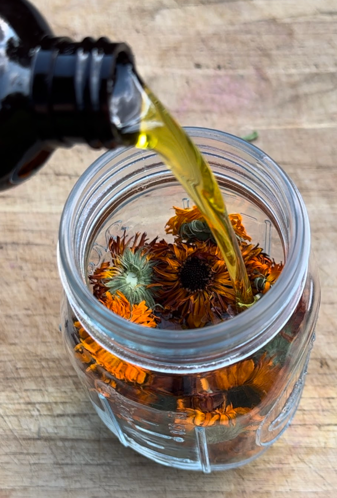 Top-down view of Calendula Body Oil being poured into a mason jar filled with dried calendula flowers on a wooden board