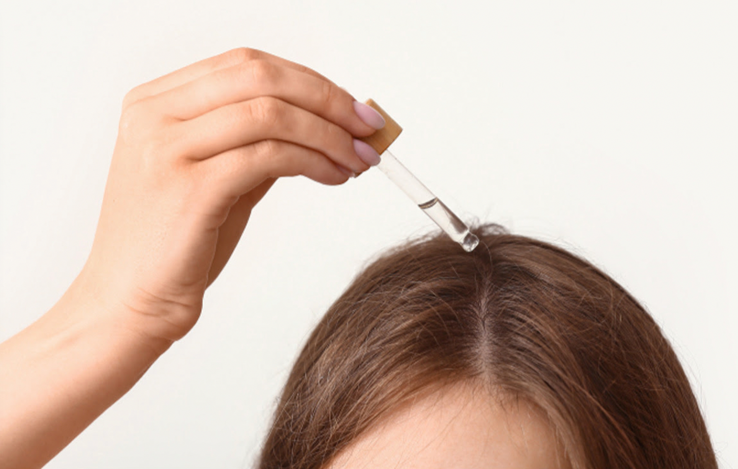 Hand applying rosemary oil to a parted brunette scalp with a glass dropper against a light background