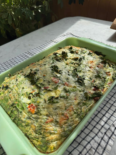 Angled top view of crustless quiche in a green baking dish showcasing spinach and tomatoes on a sunlit checkered cloth