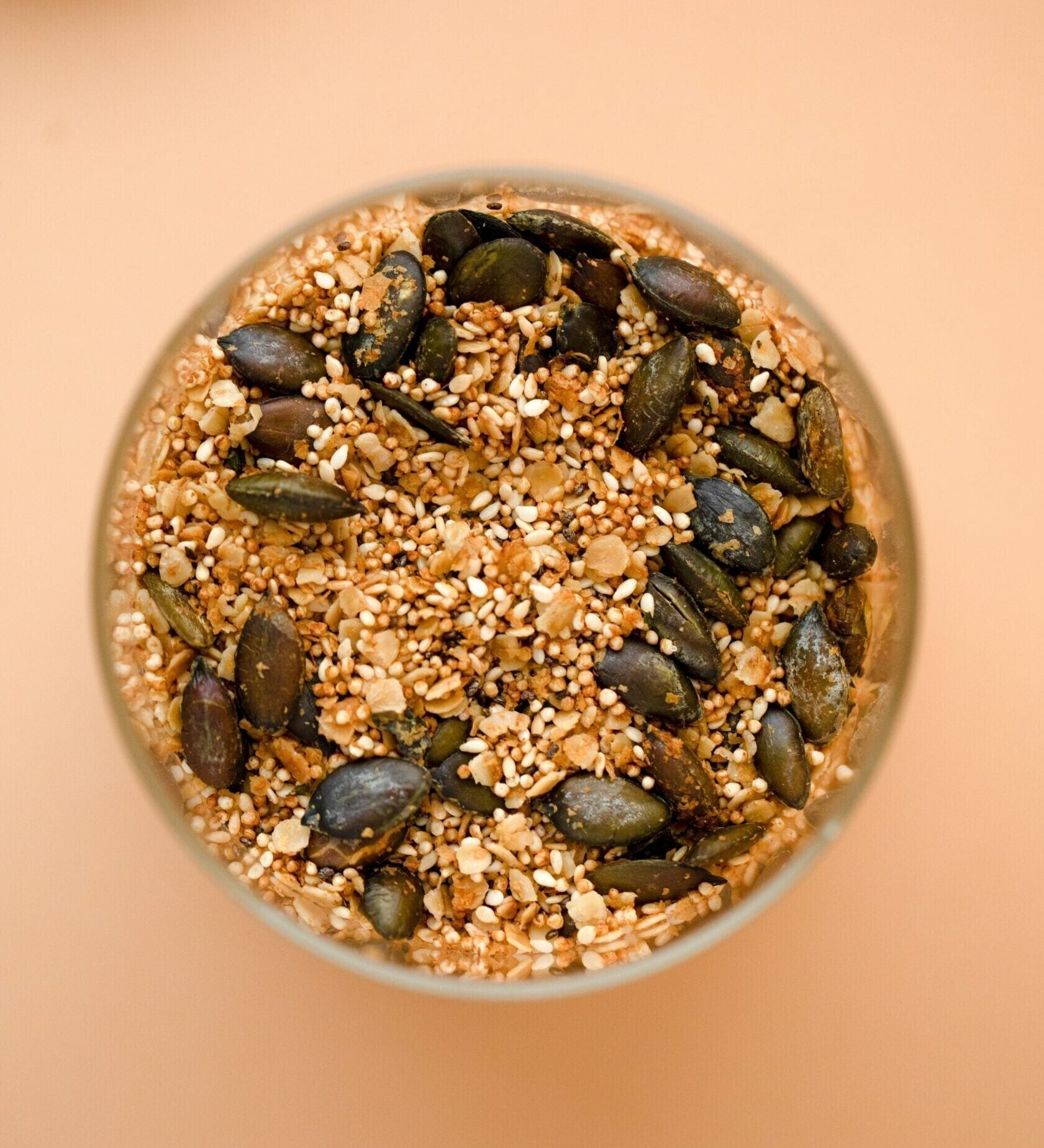 Overhead view of a jar filled with mixed seeds and grains (pumpkin seeds, sesame) on a pale peach background