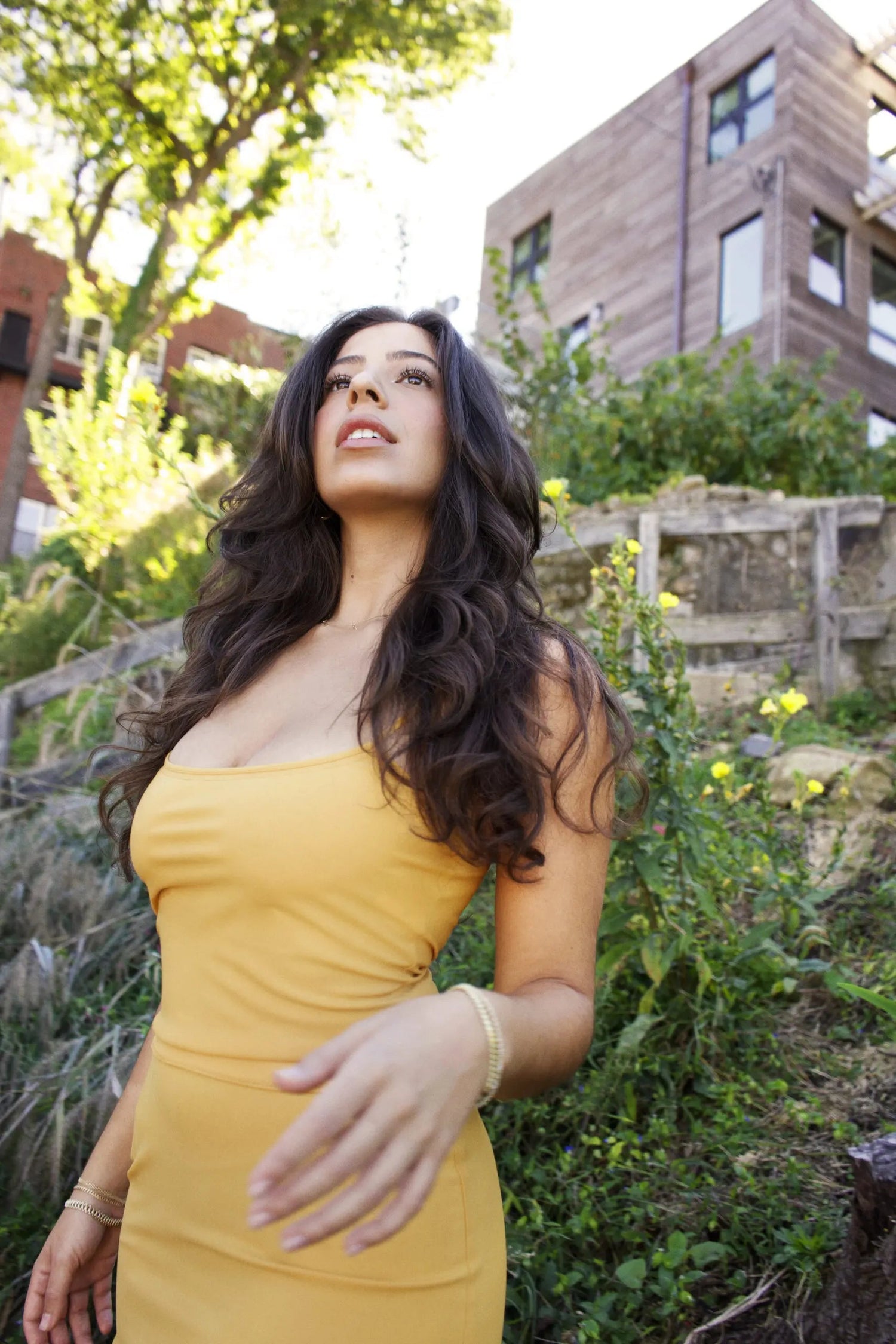 Woman in a yellow sleeveless dress looking up amid a sunlit urban garden with flowers and apartment buildings behind her