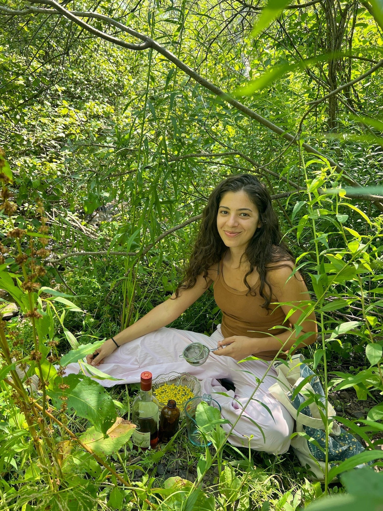 Woman preparing a flower essence in a green woodland clearing, straining yellow flower petals into a bowl with bottles nearby