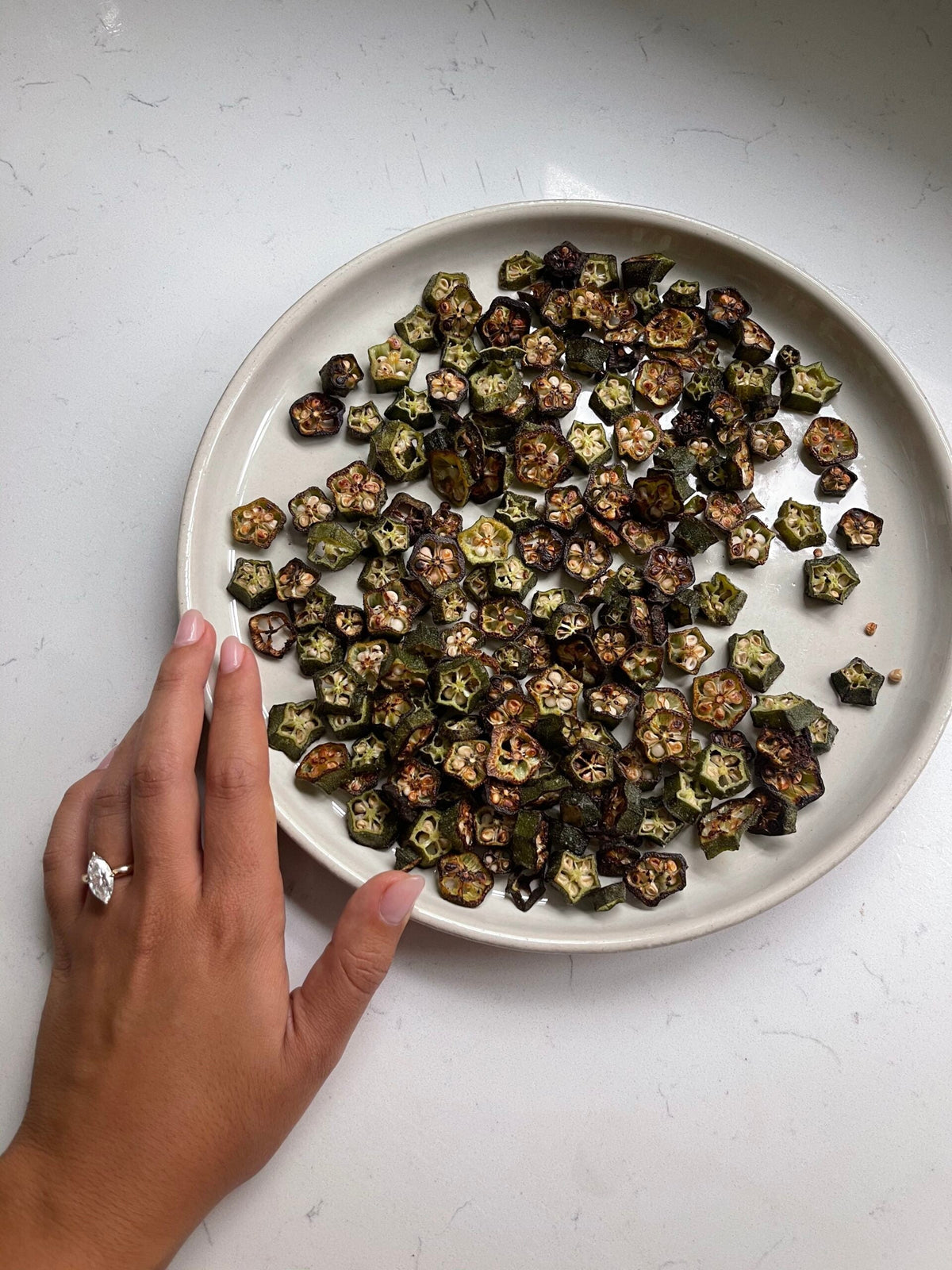 Hand beside a ceramic plate of roasted sliced okra chips on a white countertop
