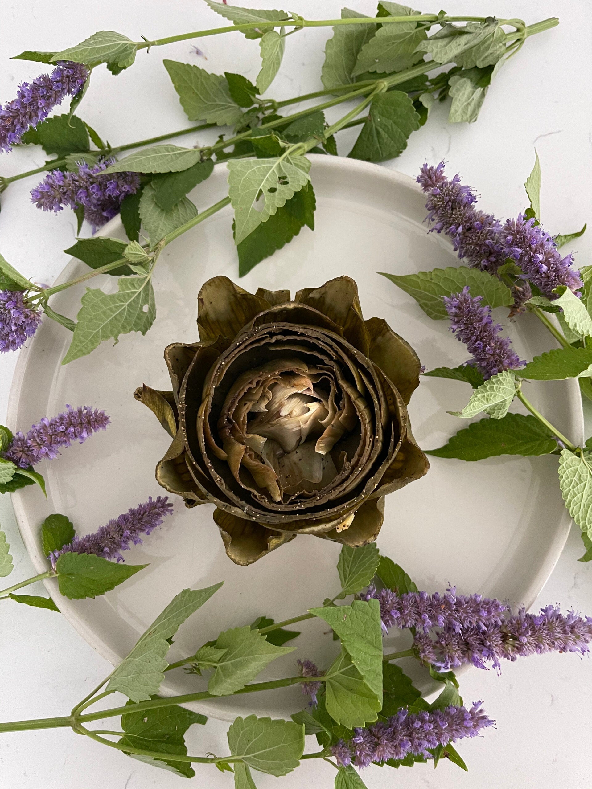 Overhead view of roasted garlic-seasoned artichoke on white plate showing opened petals and garlic flecks with purple flowers