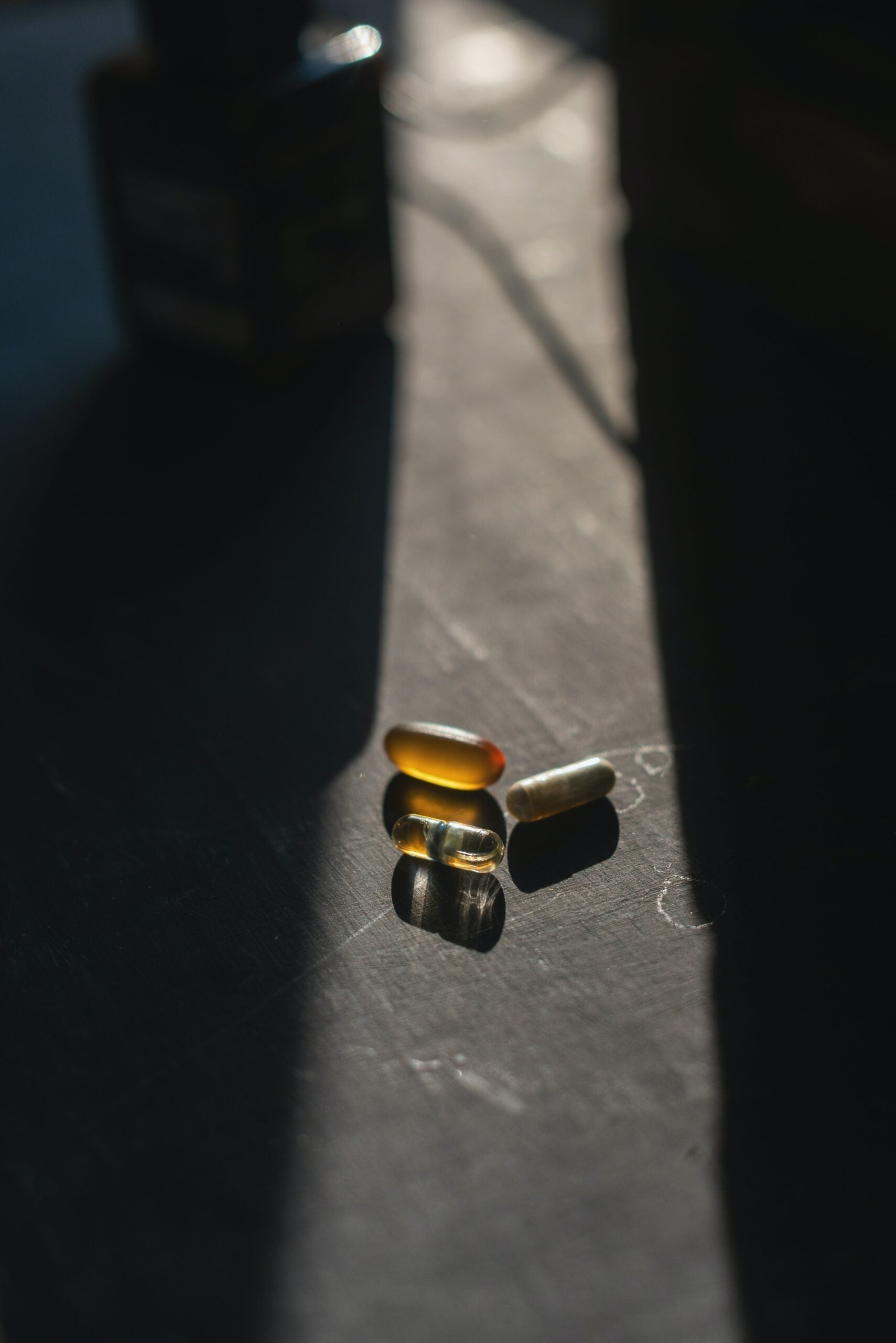 Three supplement capsules resting in a beam of sunlight on a dark tabletop