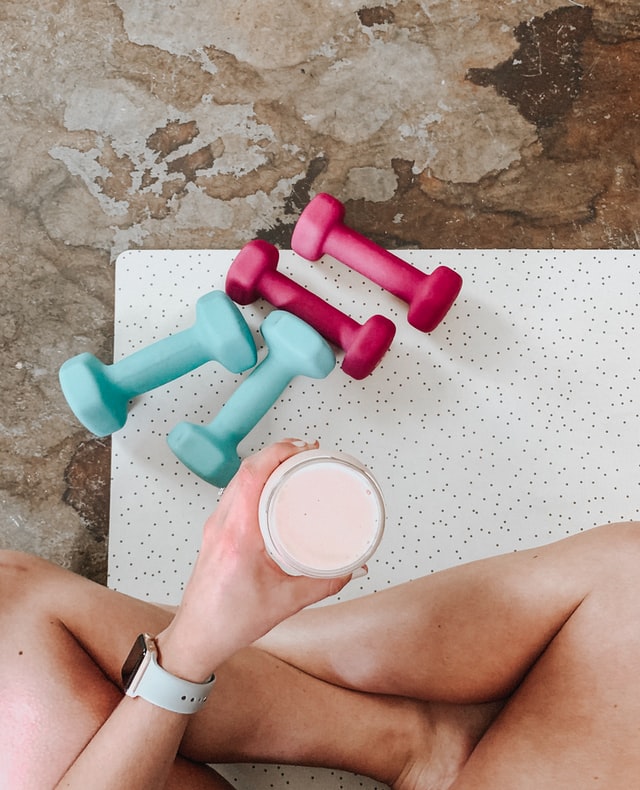 Person sitting cross-legged holding a pink drink beside colorful dumbbells on a workout mat
