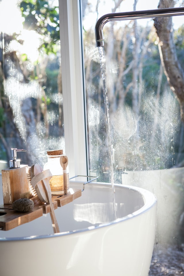 Sunlit bathtub filling from a tall faucet by a window, with a wooden bath tray holding a brush, soap dispenser, and jar