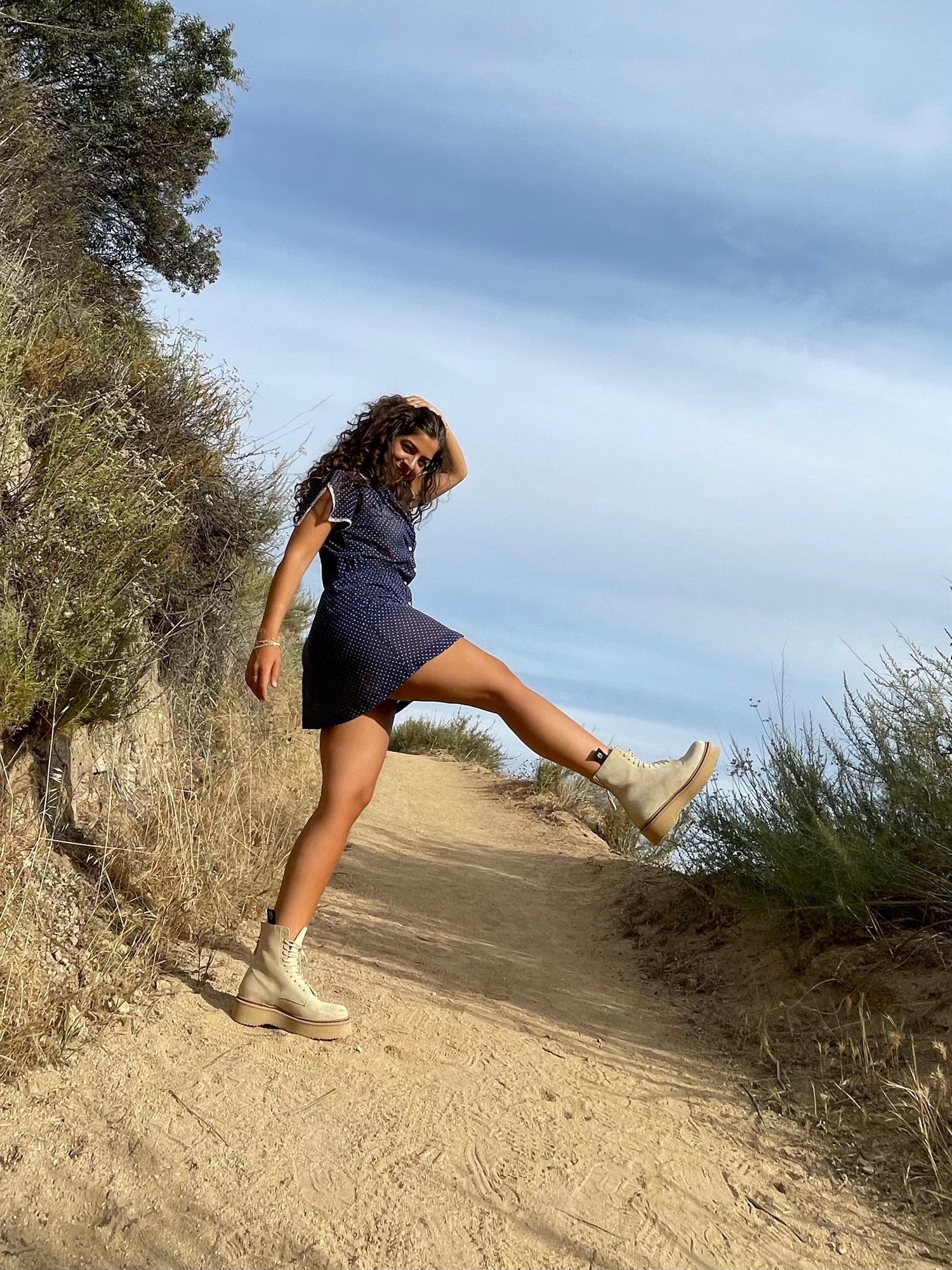 Young woman kicking her leg while walking on a sunlit dirt trail with shrubs and blue sky