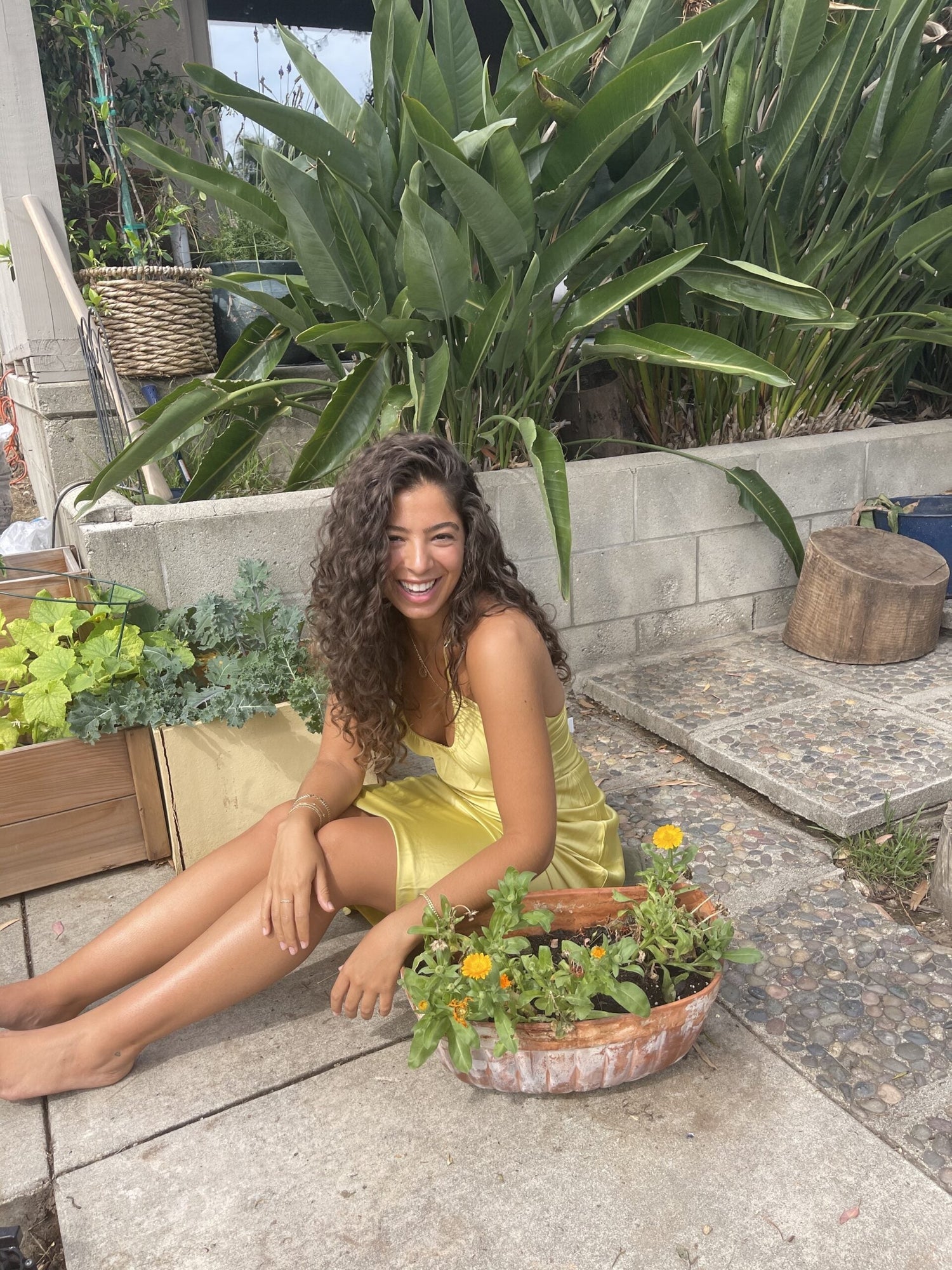 Smiling woman in a yellow dress sitting on a patio beside a planter of calendula flowers