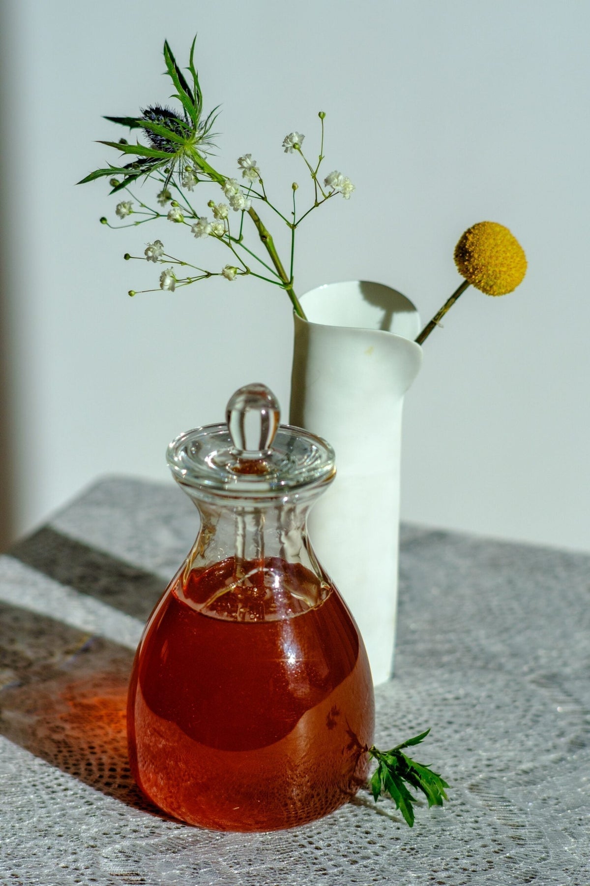 Glass carafe of amber herbal syrup beside a white ceramic pitcher with small white sprigs and a yellow billy ball on a lace-covered table