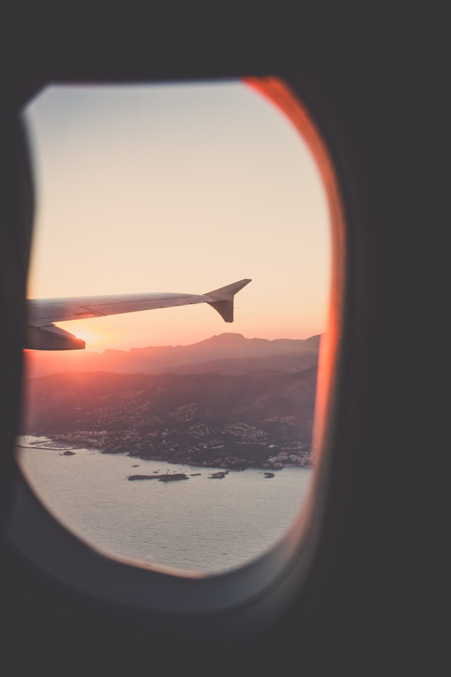 Airplane wing flying over coastal mountains at sunset seen through a passenger window