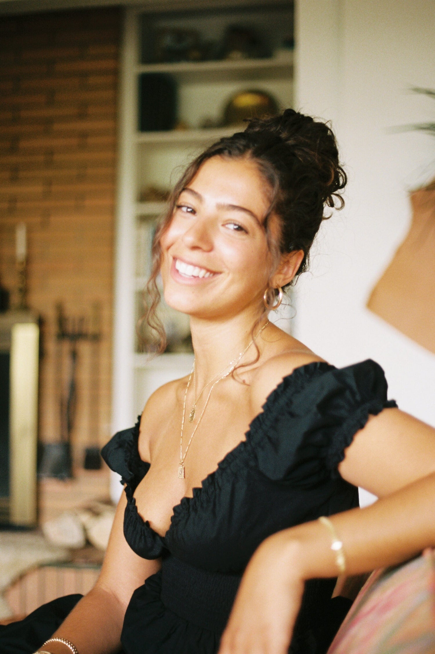 Young woman smiling seated in a cozy living room wearing a black off-the-shoulder top, layered gold necklaces and hoop earrings