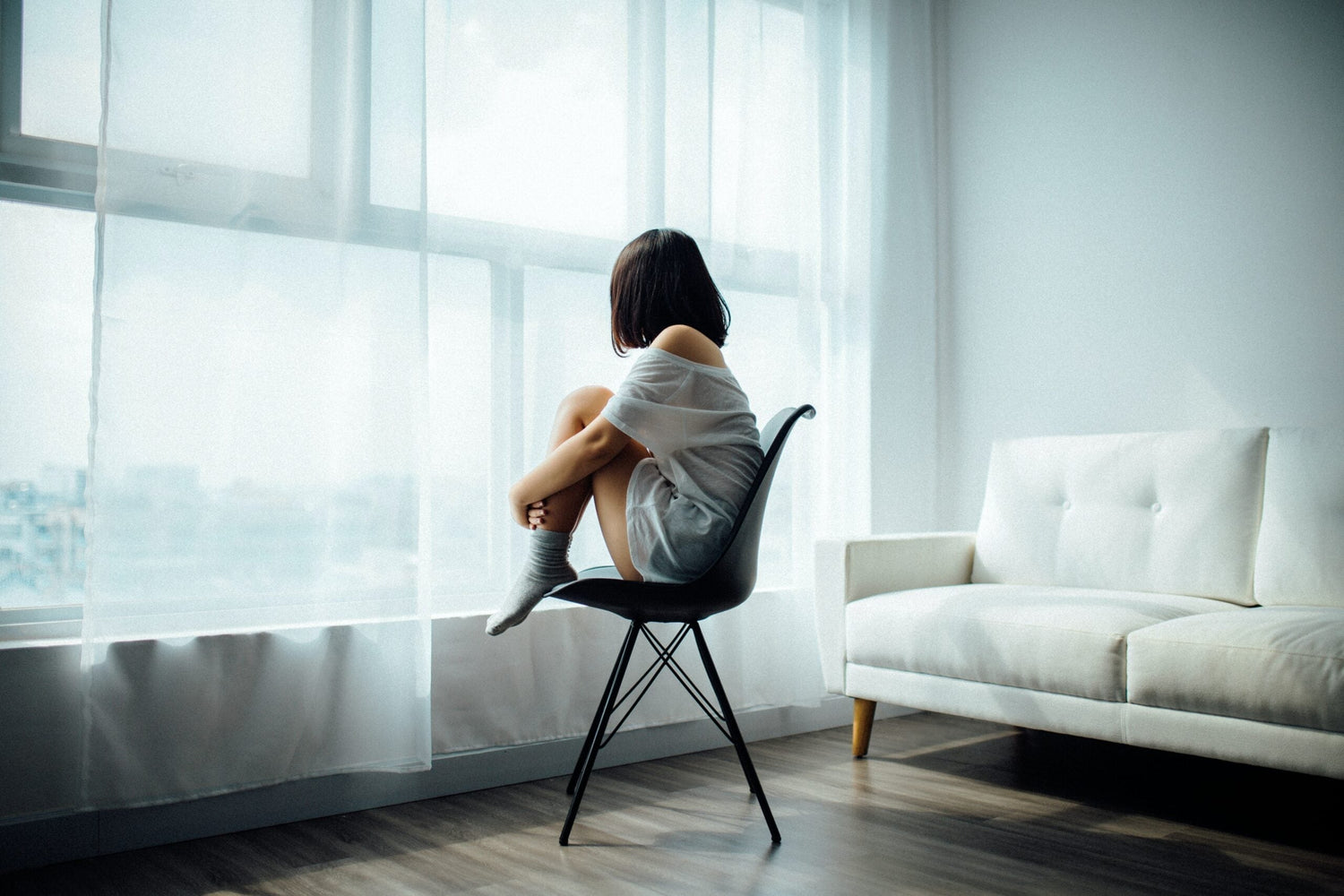 Woman curled on a chair by a large sunlit window in a minimal living room, looking outside