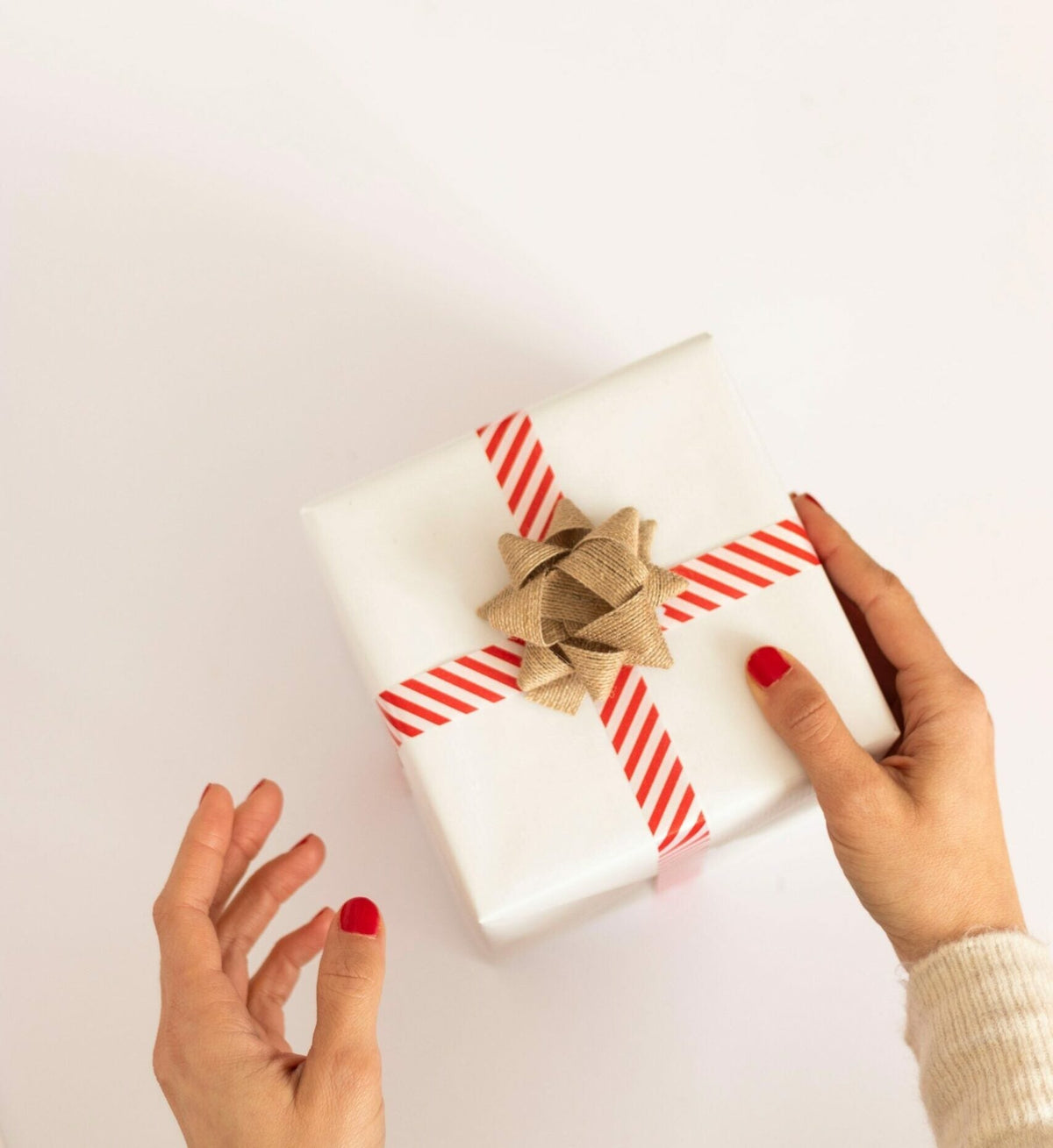 Hands holding a wrapped white gift box with red striped ribbon and gold bow on a white background