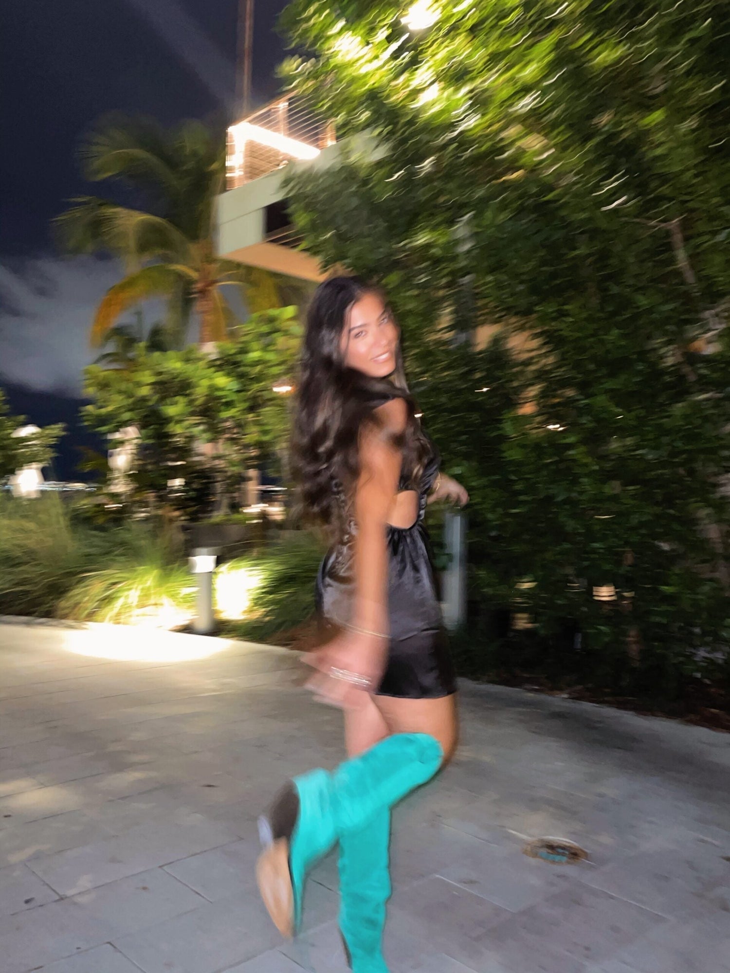Woman in black satin dress turning and smiling at night by lit plants, wearing teal knee-high boots
