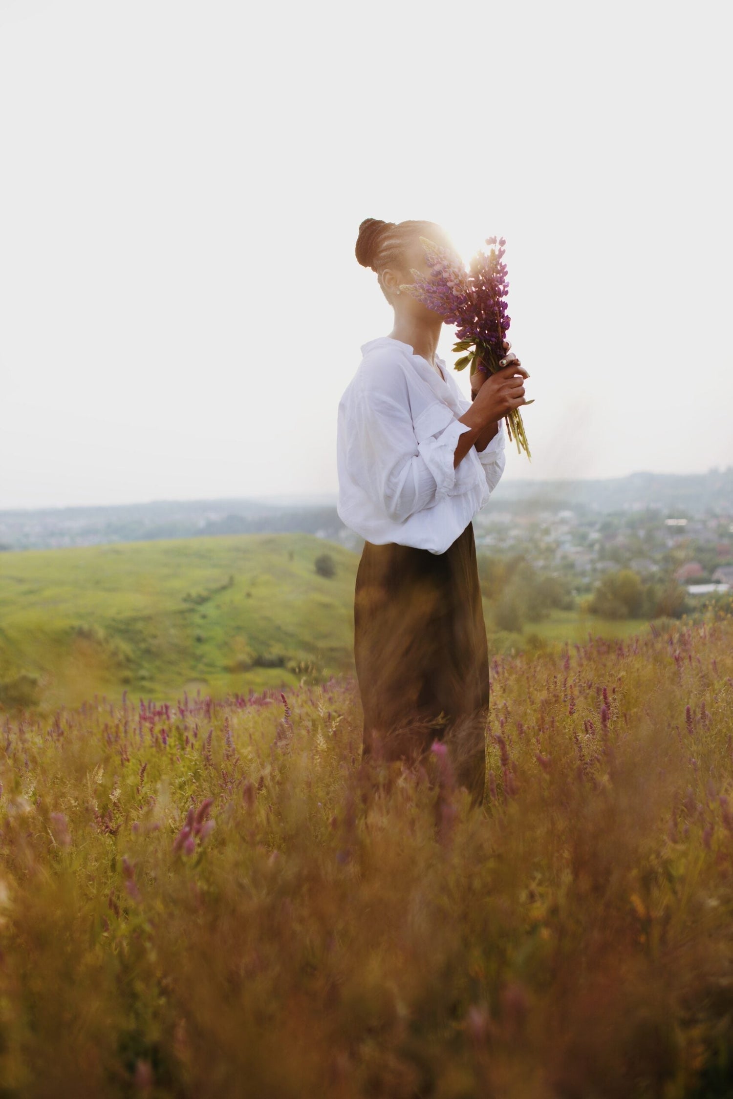 Woman in a white blouse holding a bouquet of purple wildflowers in a sunlit meadow with rolling hills in the background