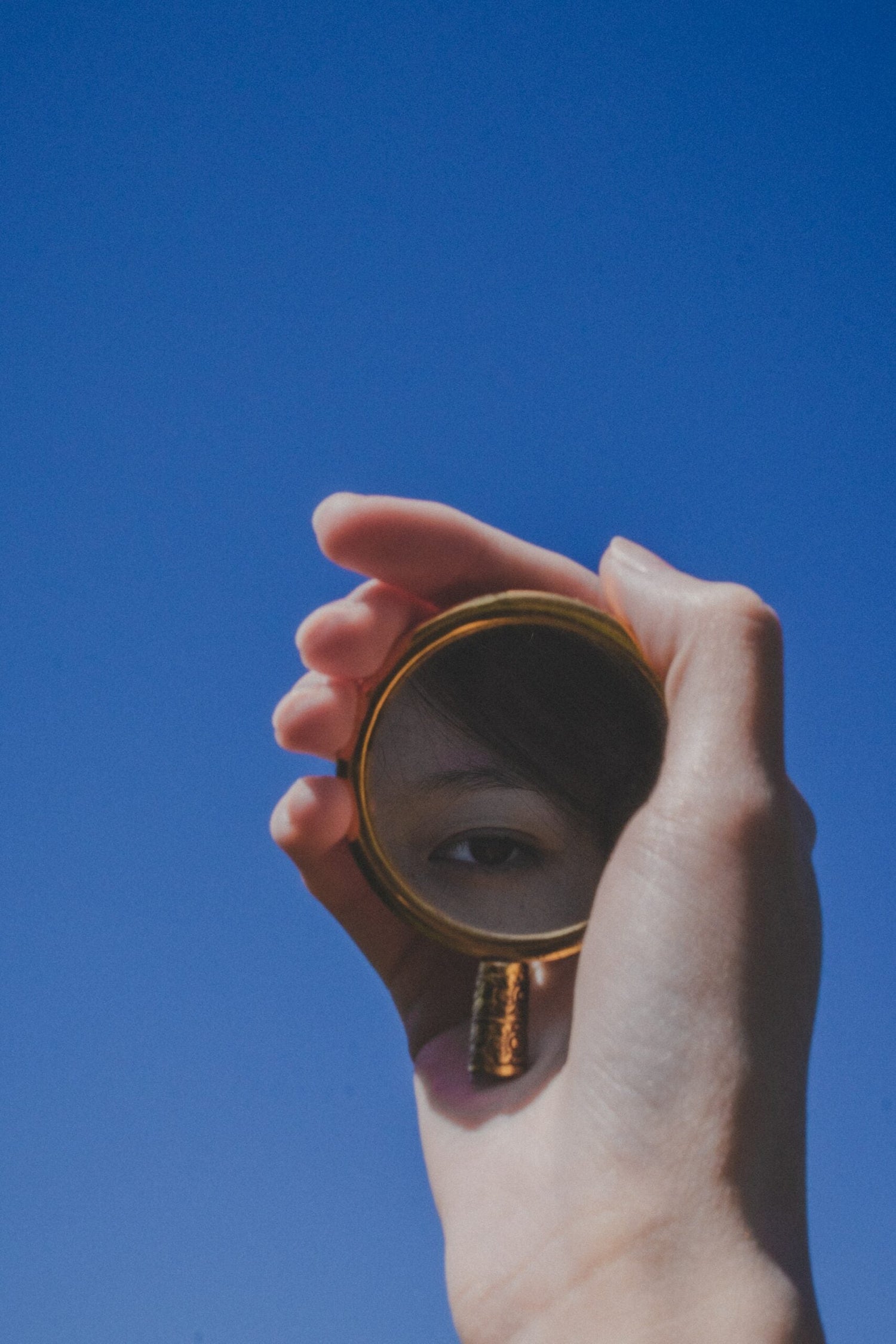 Hand holding a small round mirror reflecting one eye against a clear blue sky