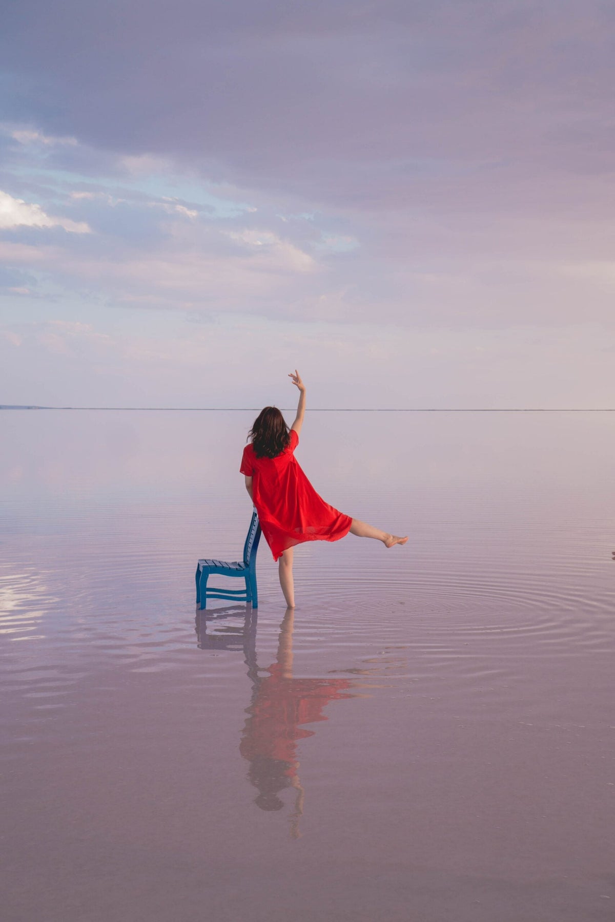 Woman in a red dress balances beside a chair in shallow water beneath a pastel sky