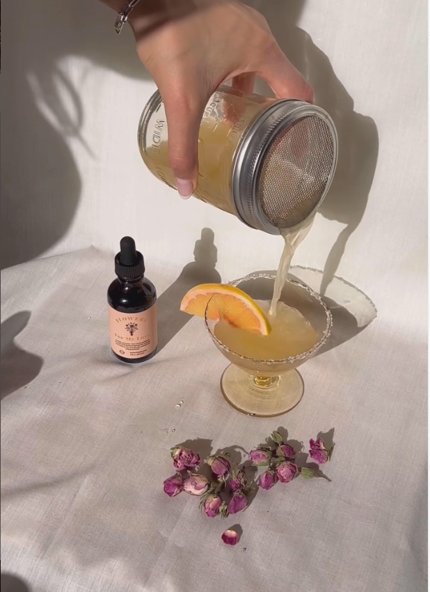 Hand pouring rose mezcal through a mesh strainer into a sugar-rimmed coupe with a grapefruit slice and dried rosebuds on white linen