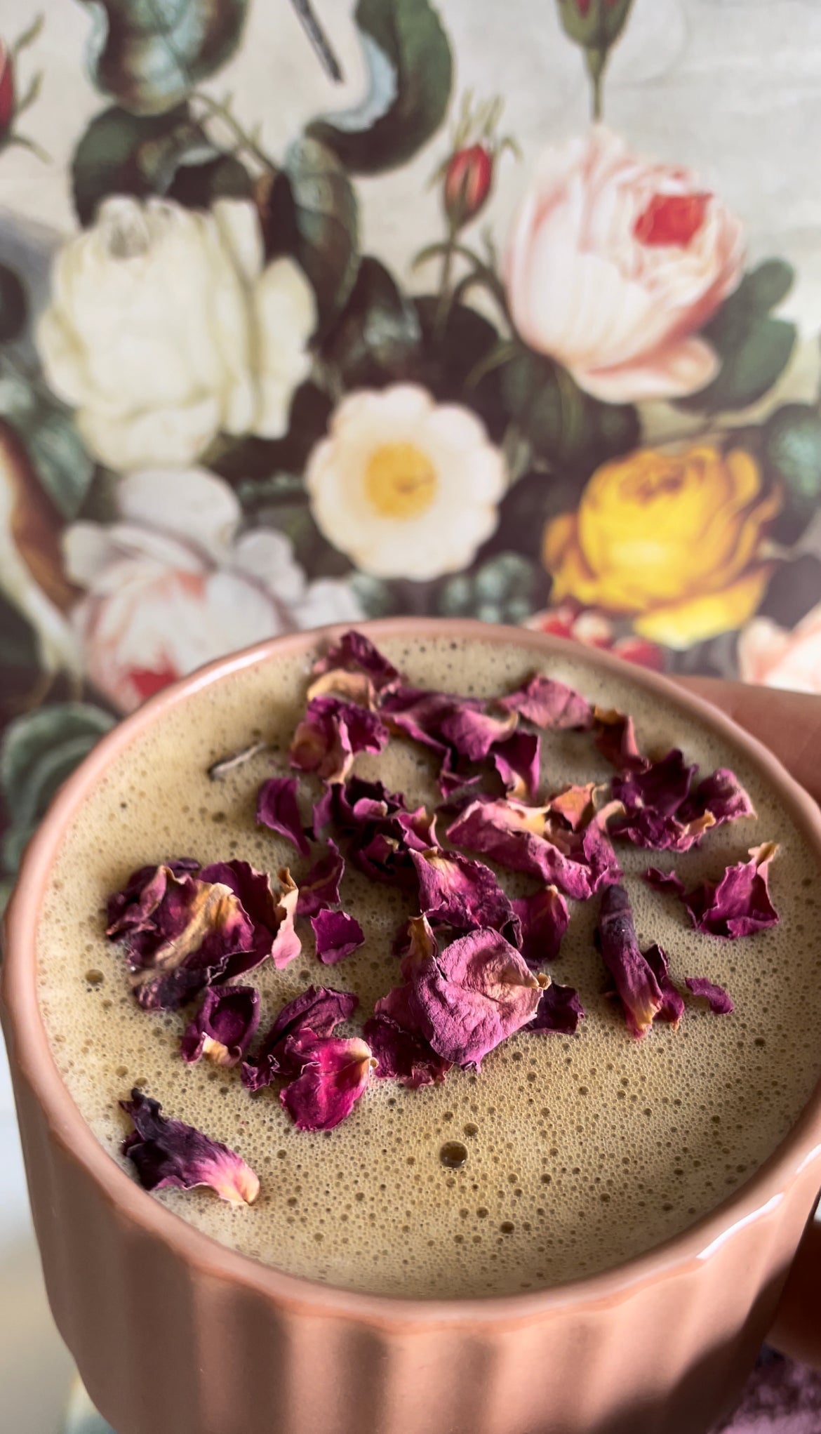 Close-up of a rose cardamom latte in a pink ceramic mug topped with dried rose petals against a vintage floral backdrop