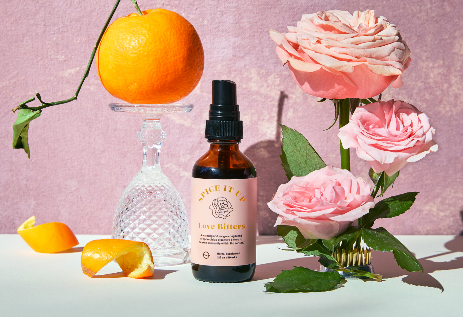 Front of Love Bitters amber spray bottle with blush label and pump, with an orange on crystal and roses on pink backdrop