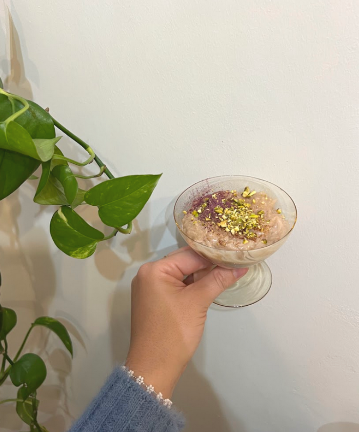 Hand holding a glass of rose rice pudding topped with crushed pistachios and rose powder against a white wall with a trailing pothos plant